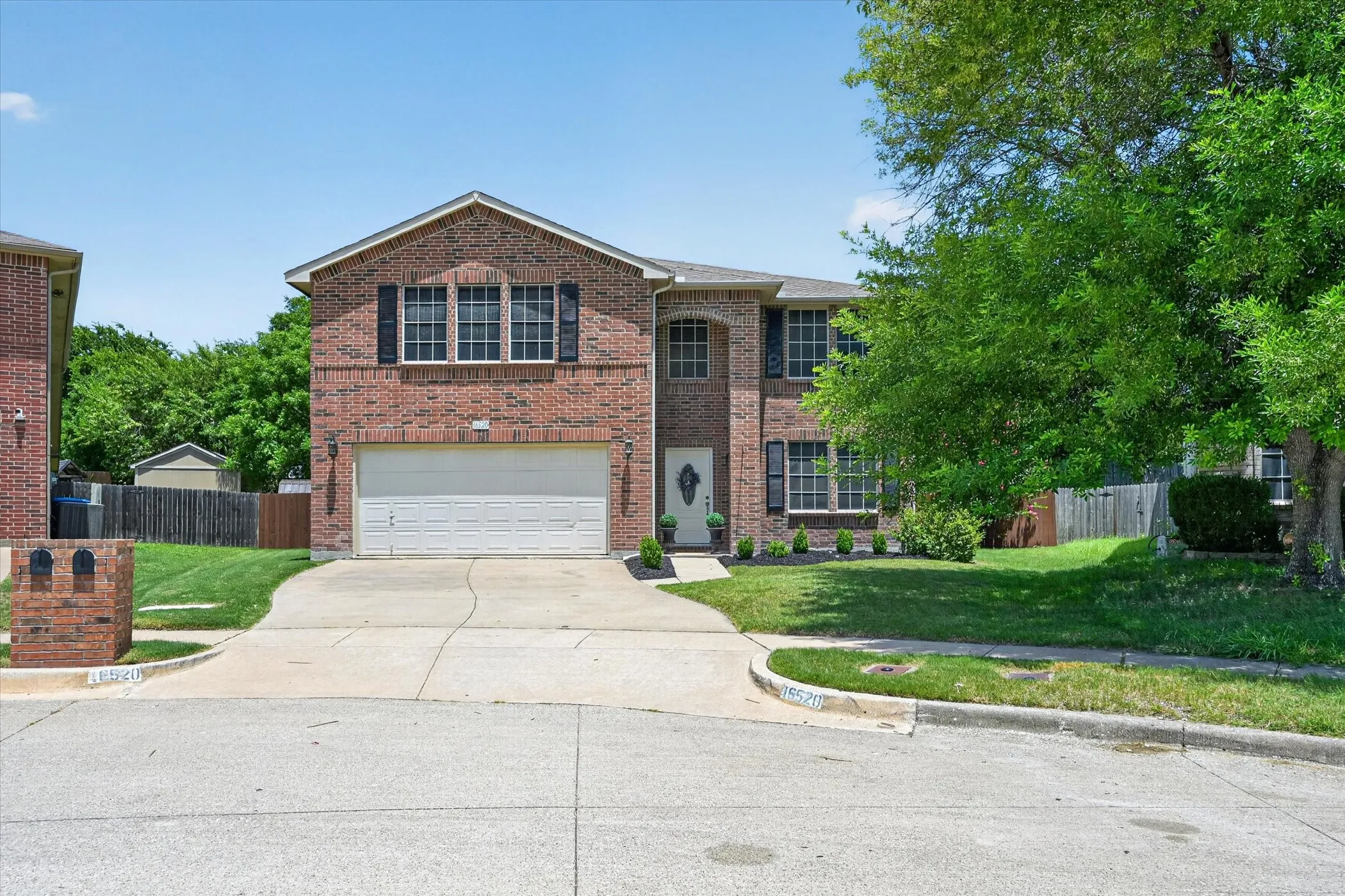 Brick facade residence featuring a two-car garage, mature front yard landscaping, concrete driveway, and a decorative front entry door