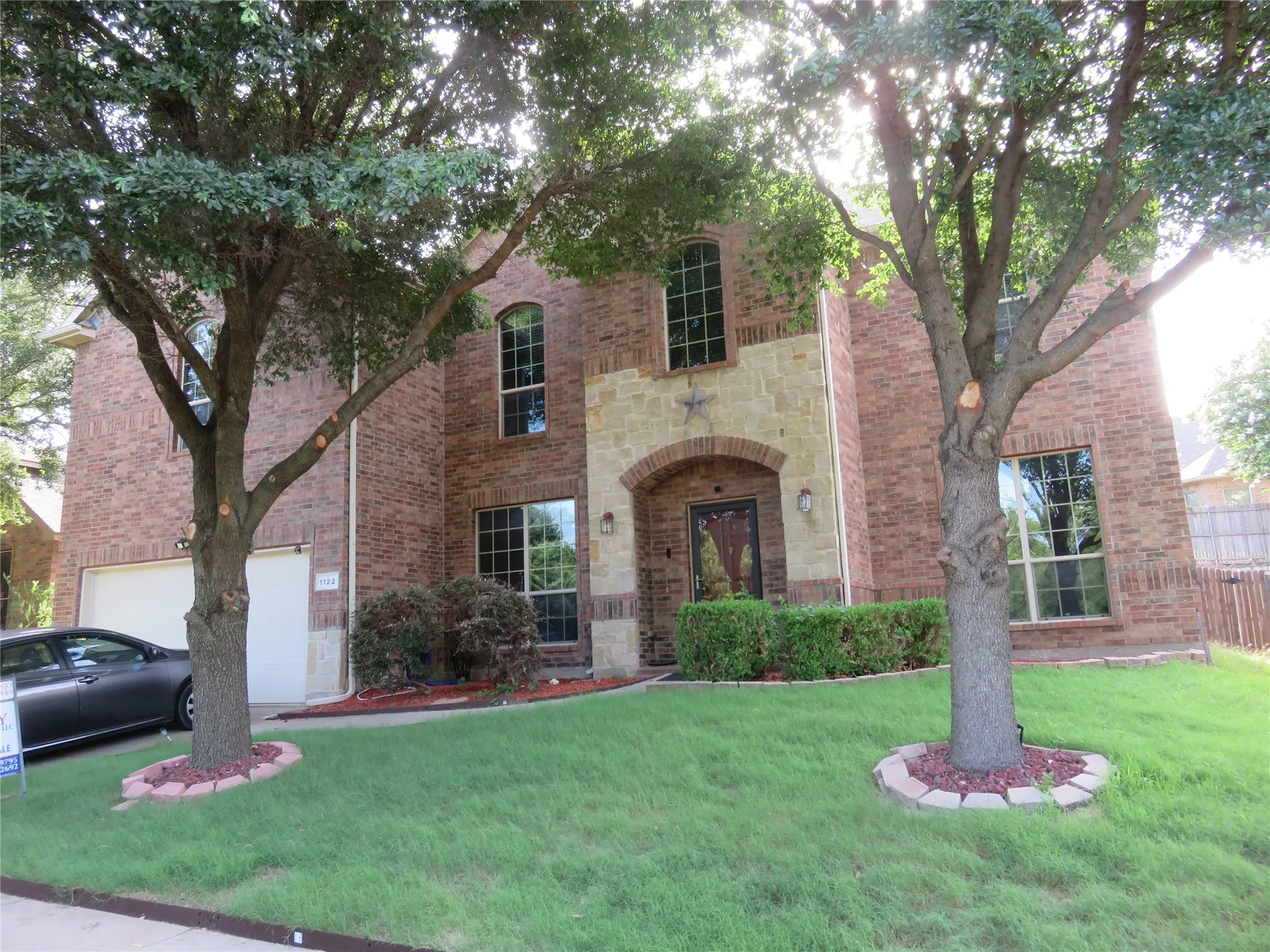 Brick and stone facade featuring an arched entryway, two prominent trees, a two-car garage, and established landscaping