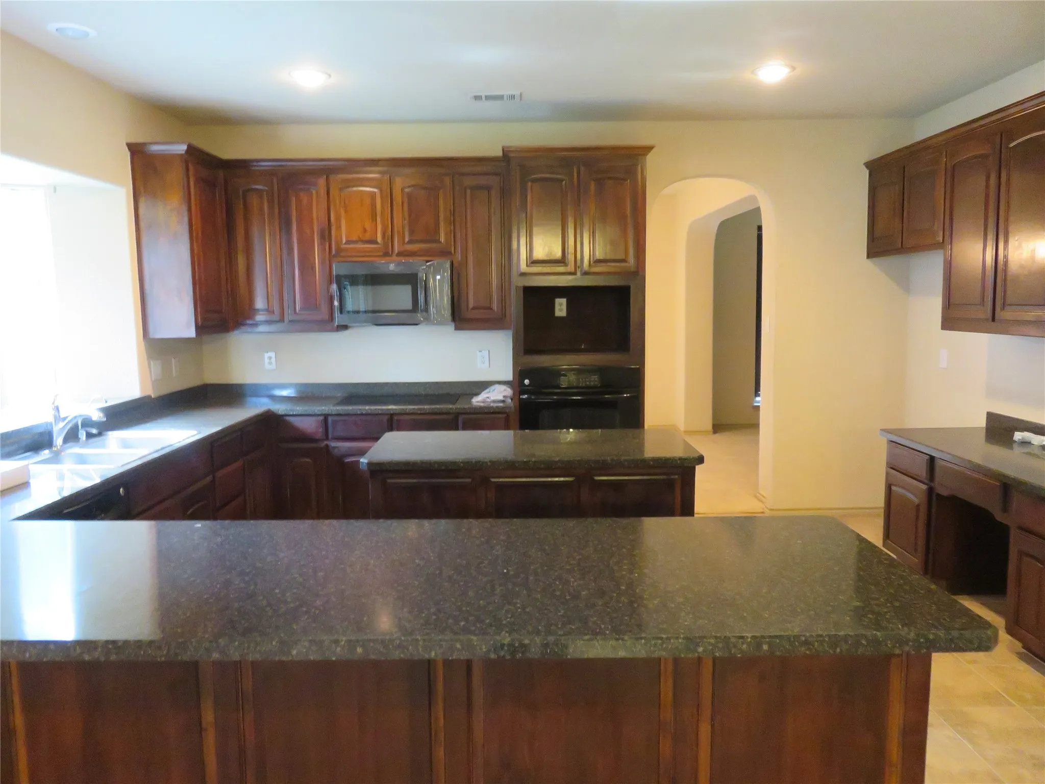Kitchen featuring dark wood cabinetry, speckled countertops, a double basin sink, built-in microwave, and arched doorway