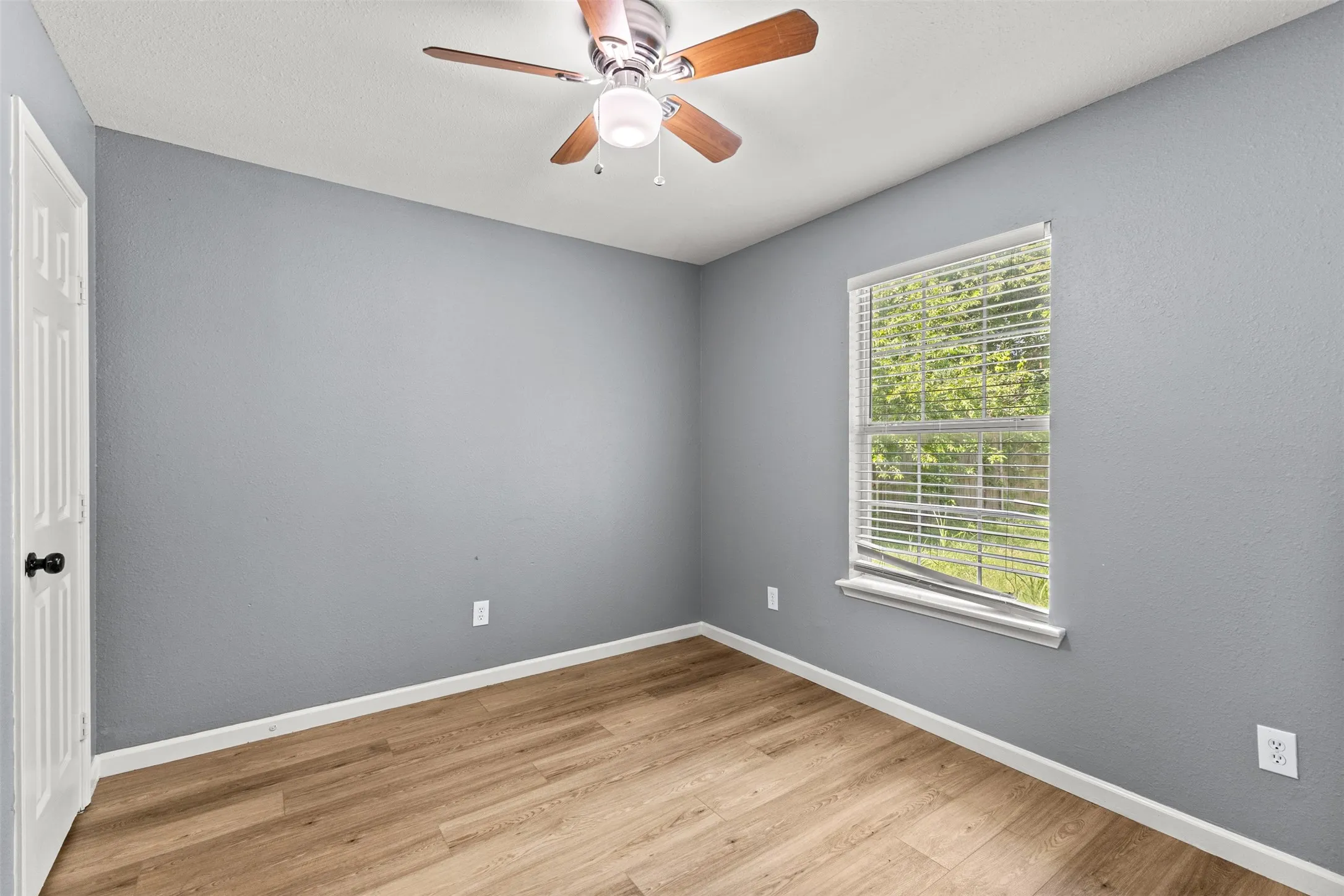 Room featuring wood-finish flooring, a ceiling fan with integrated lighting, a window with horizontal blinds, and a white paneled door with a black knob