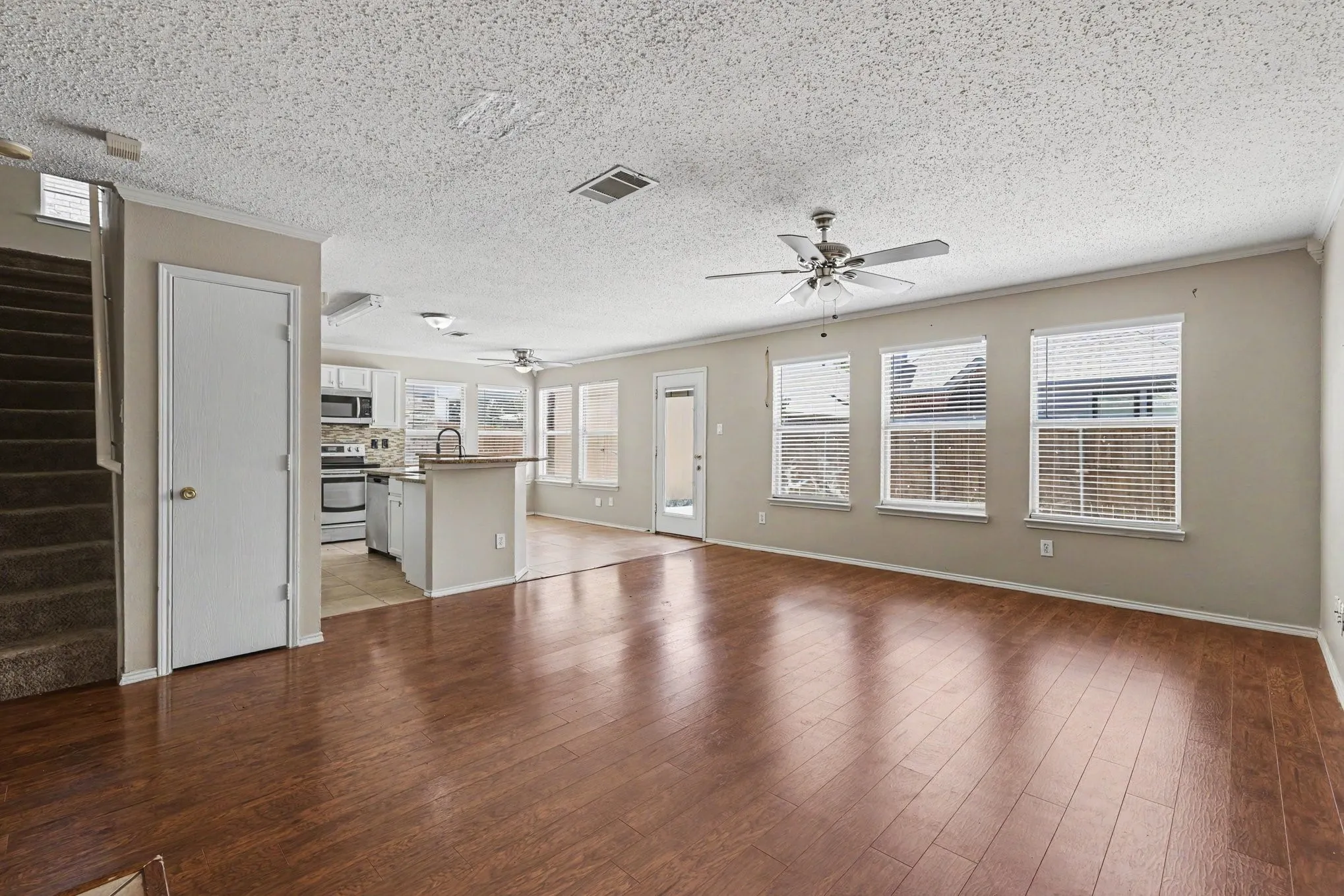 Spacious living area featuring wood-finish flooring, neutral wall tones, and multiple windows with blinds