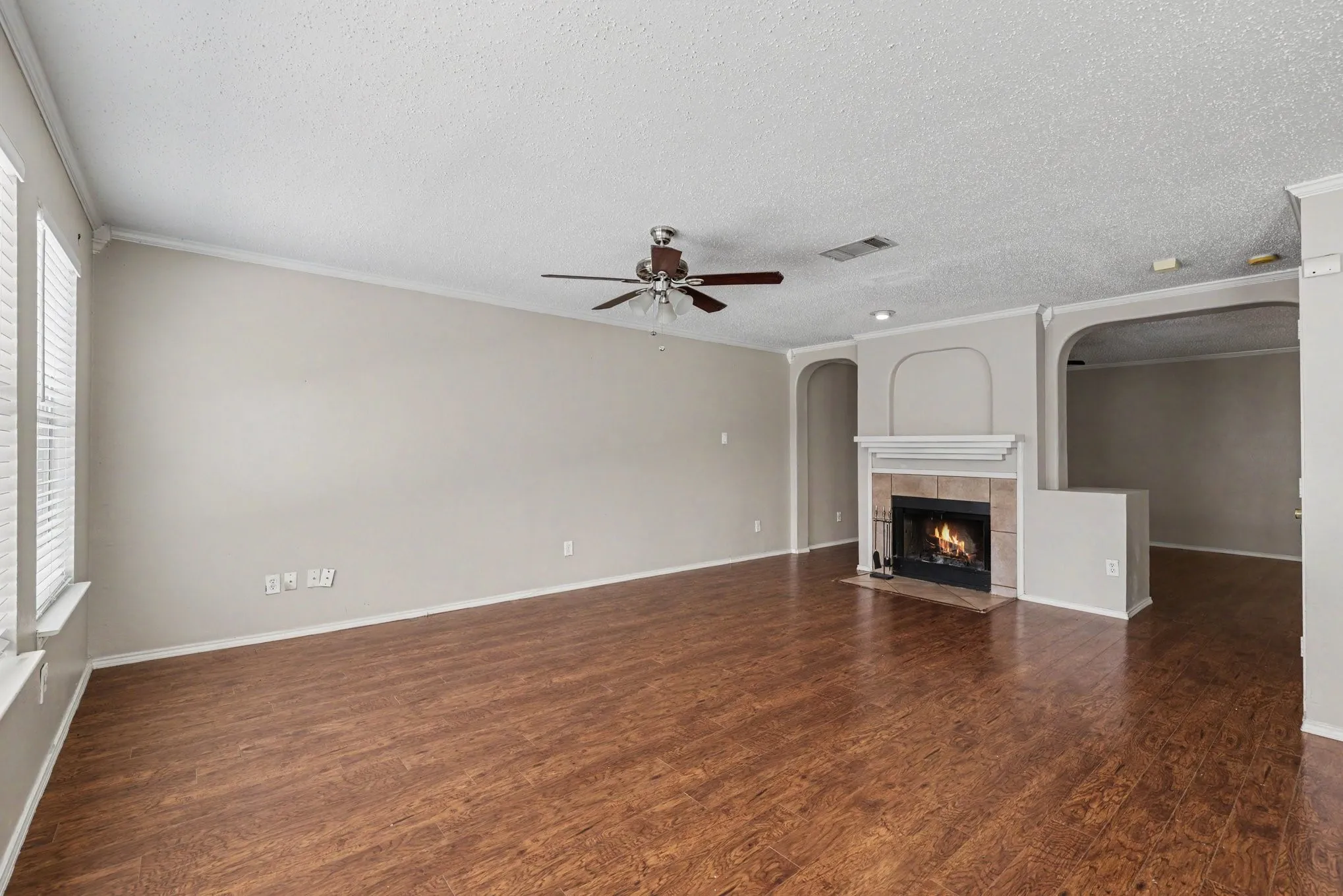 Spacious living area featuring wood-finish flooring, a tiled fireplace with mantel, ceiling fan, and crown molding