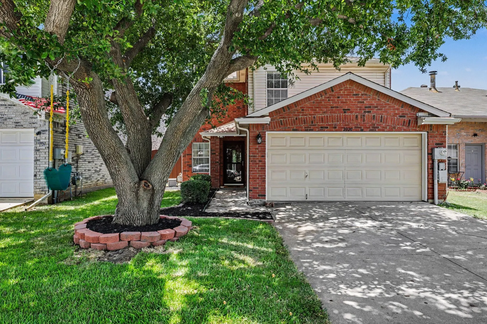 Brick facade with an attached two-car garage, concrete driveway, and mature front yard tree with a brick-edged planting bed