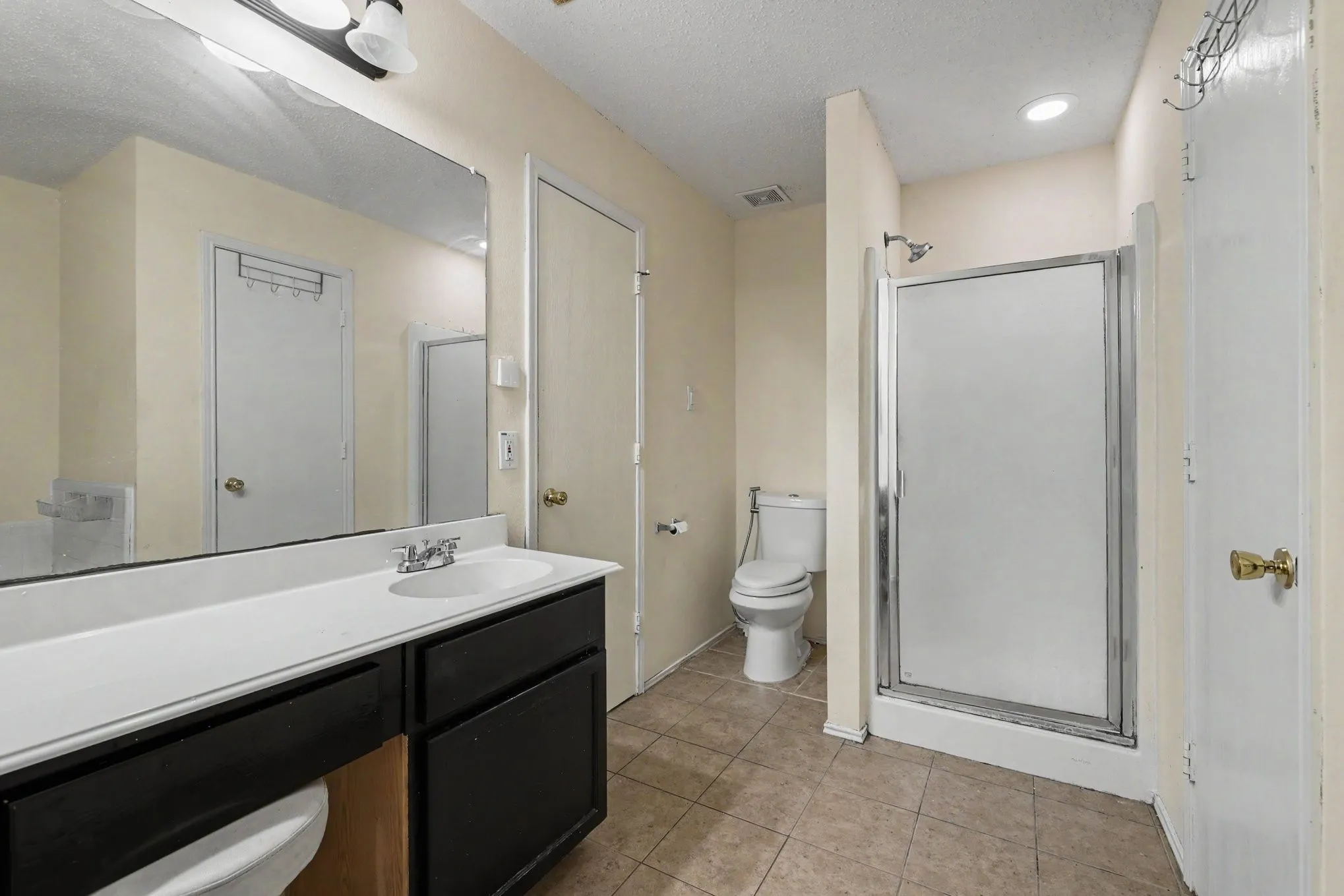 Bathroom featuring a single vanity with dark cabinetry and a white countertop, large wall-mounted mirror, stand-up shower with a frosted glass enclosure, and neutral-toned floor tiling