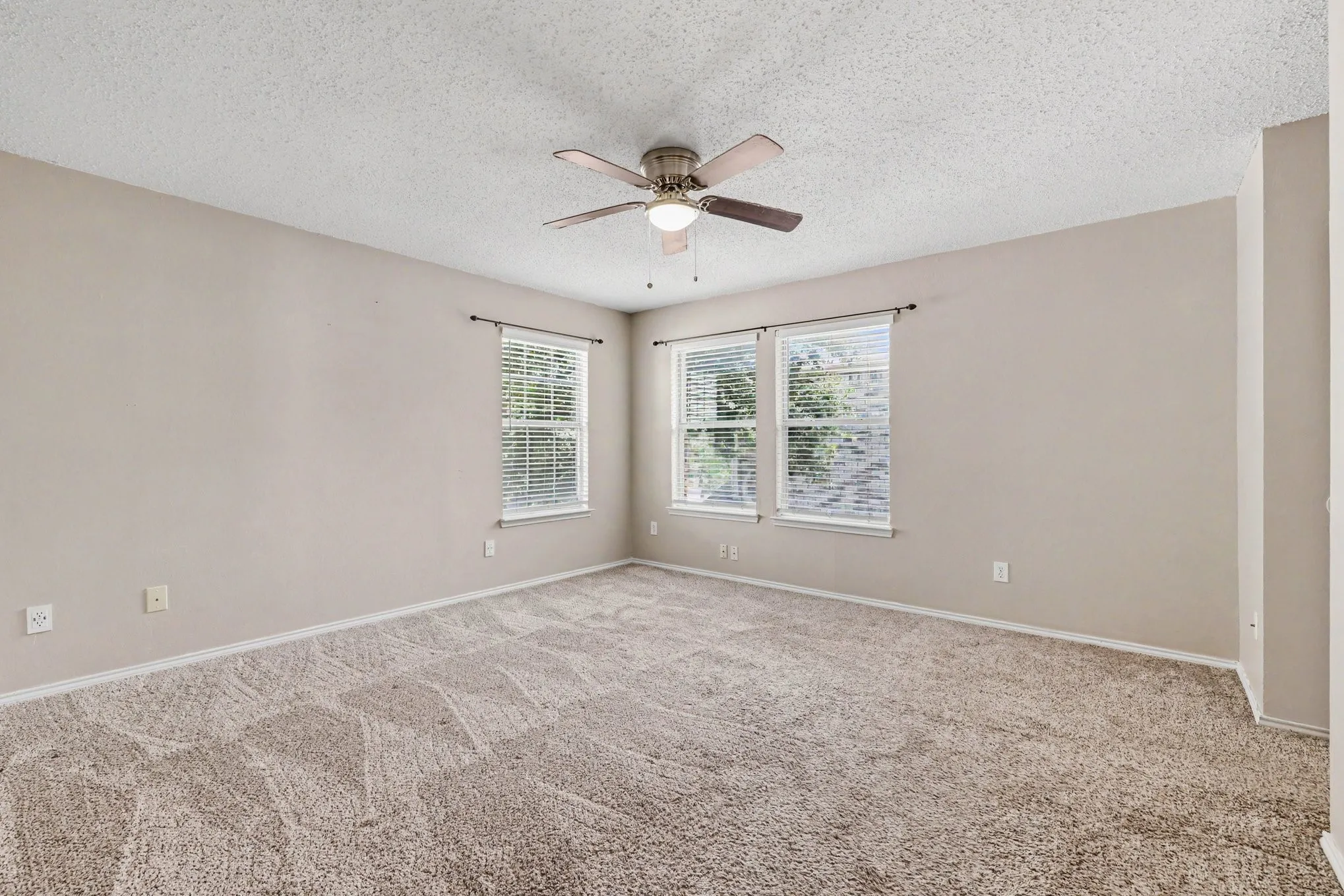 Spacious room featuring textured carpeting, multiple windows with blinds, a ceiling fan with integrated lighting, neutral wall paint, and white trim