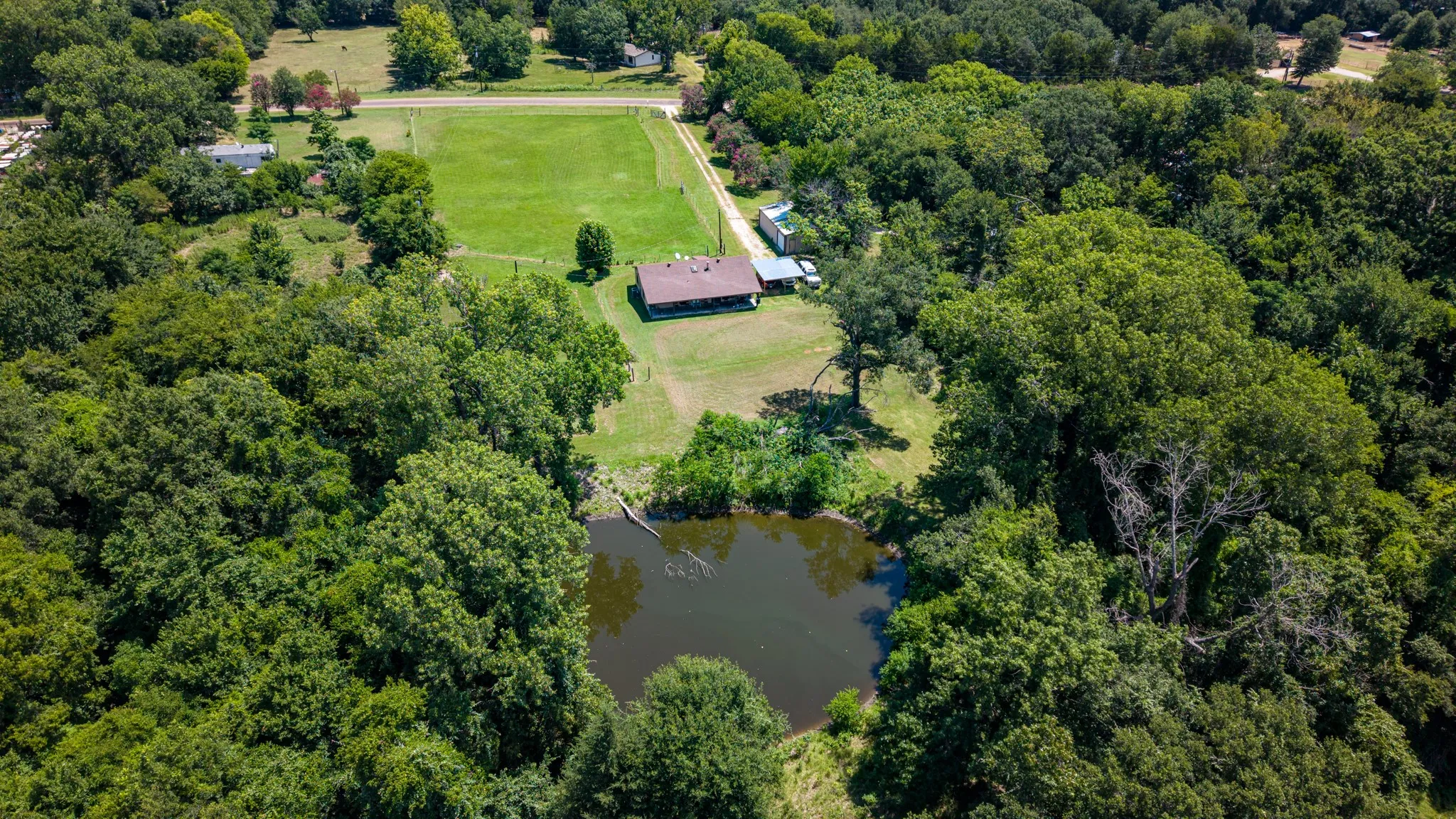 Expansive aerial perspective showcasing a property surrounded by mature trees, featuring a private pond, a residential structure with a brown roof, an adjacent outbuilding, and a cleared grassy area