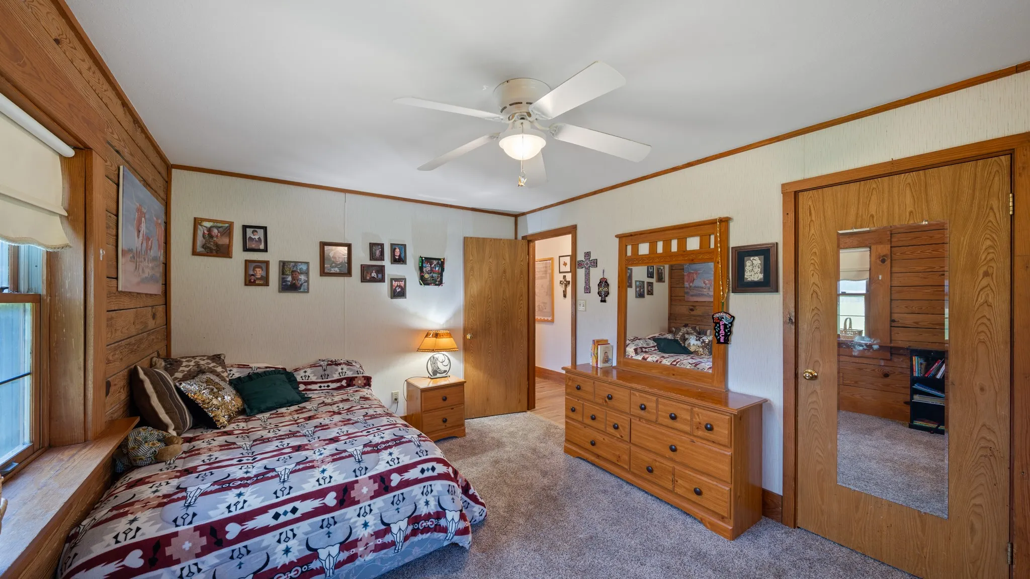 Bedroom featuring wood-paneled accent wall, a window with a built-in ledge, light-toned carpeting, a ceiling fan, and solid wood trim