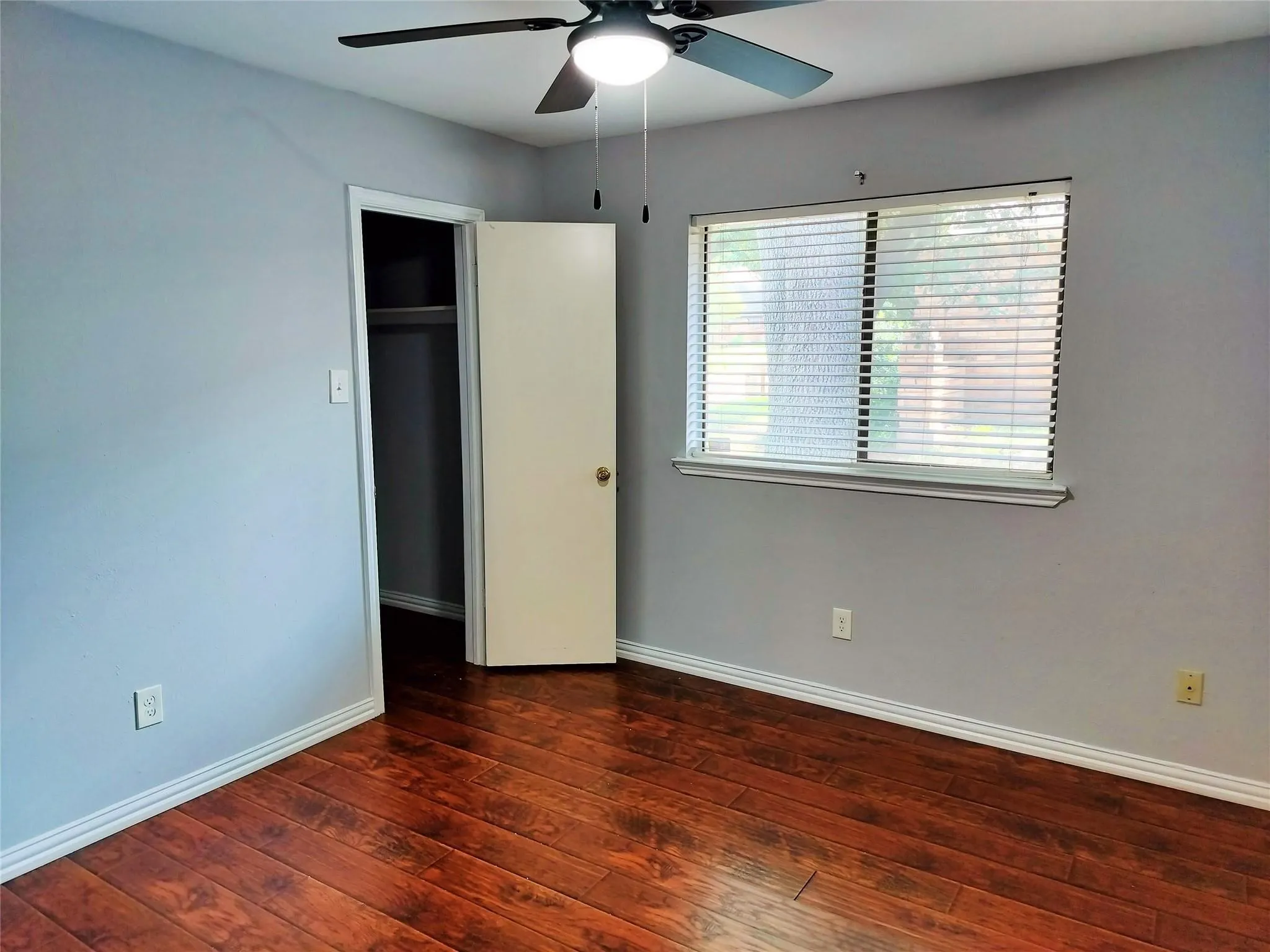 Room with wood-finish flooring, a ceiling fan with integrated lighting, and a window featuring horizontal blinds