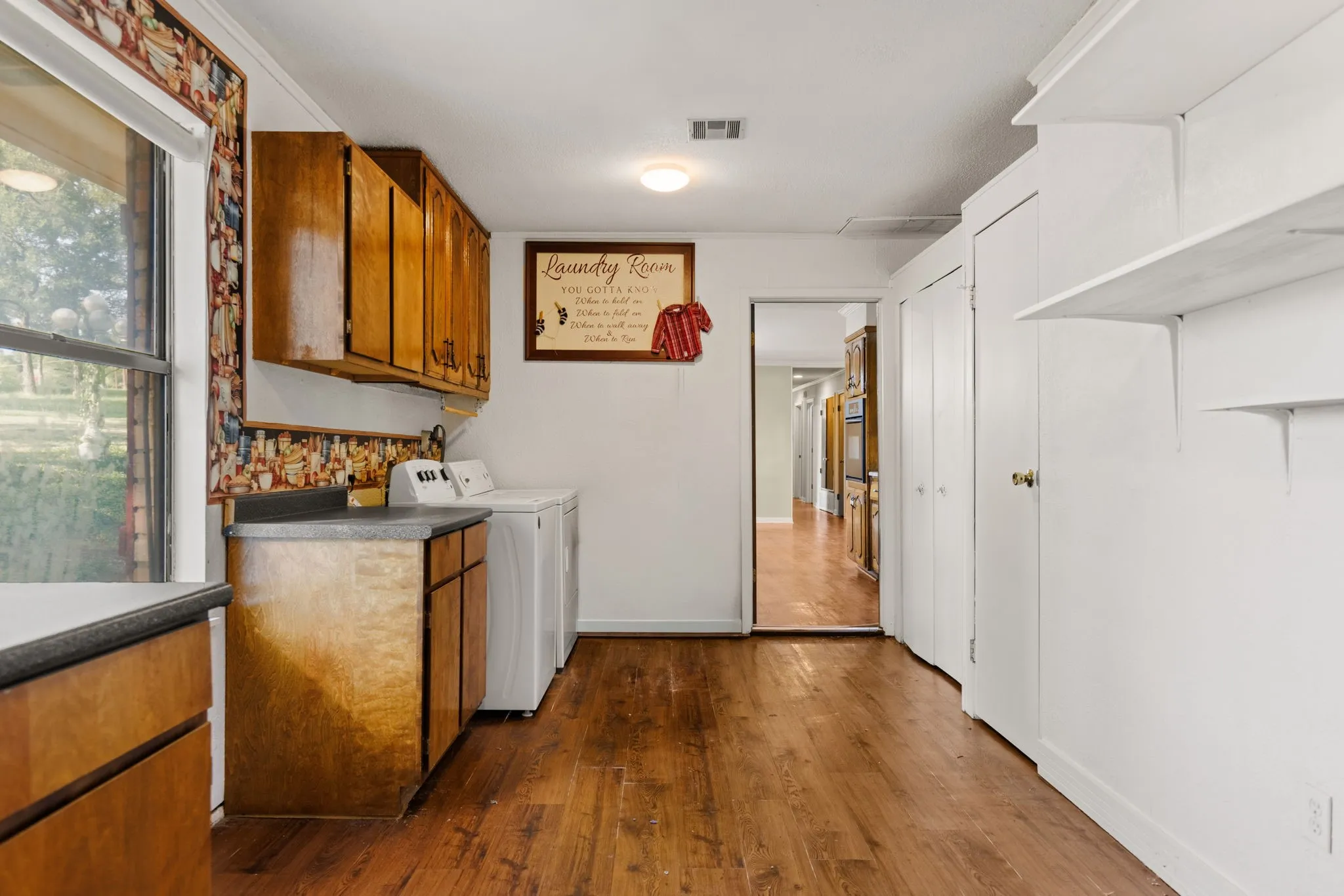Utility area featuring wood-finish flooring, a window, wood cabinetry, a countertop, and wall-mounted shelving