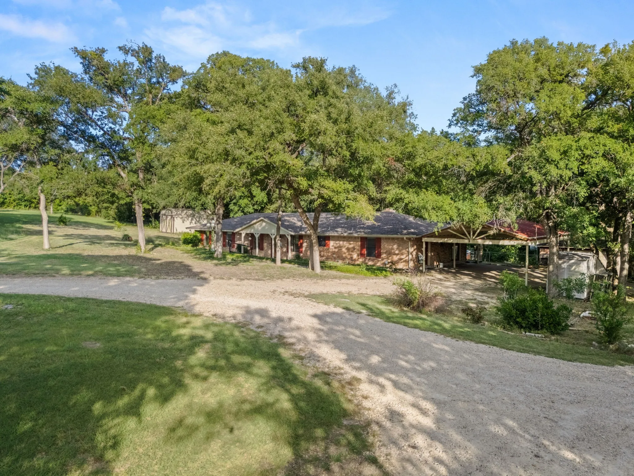 Spacious property featuring a gravel driveway, mature trees, a brick exterior, an attached covered carport, and a detached shed