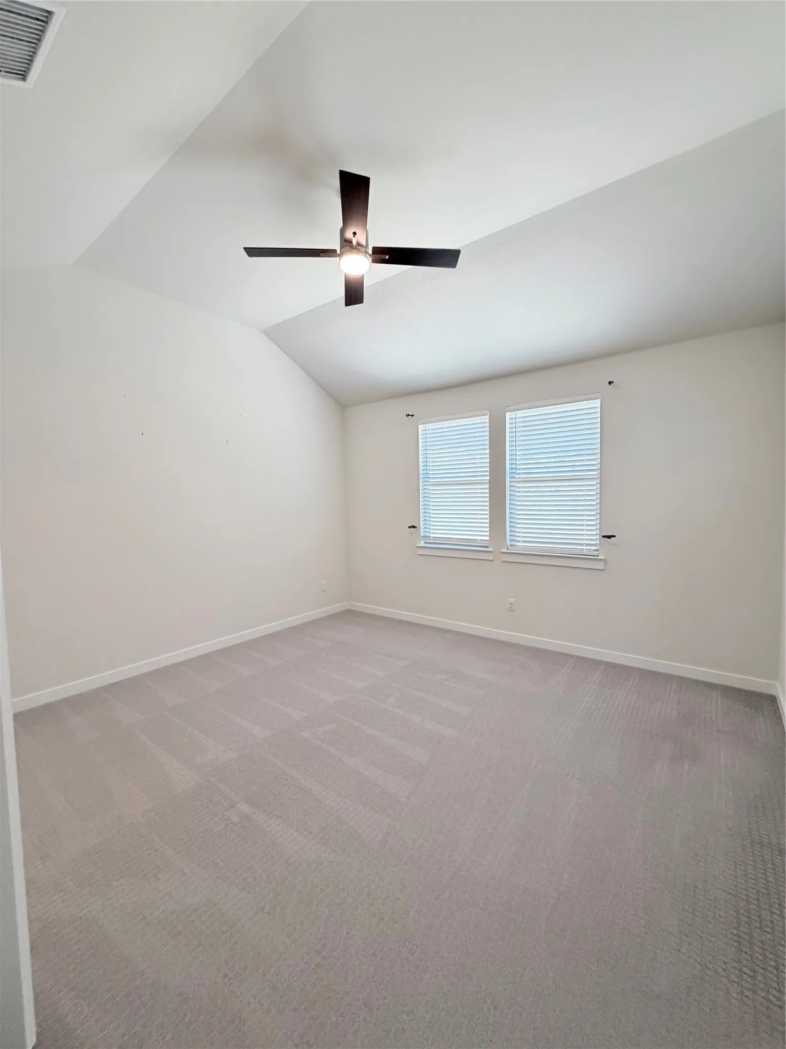 Spacious room featuring vaulted ceilings, light-colored walls, gray carpeting, a contemporary ceiling fan, and dual windows with blinds