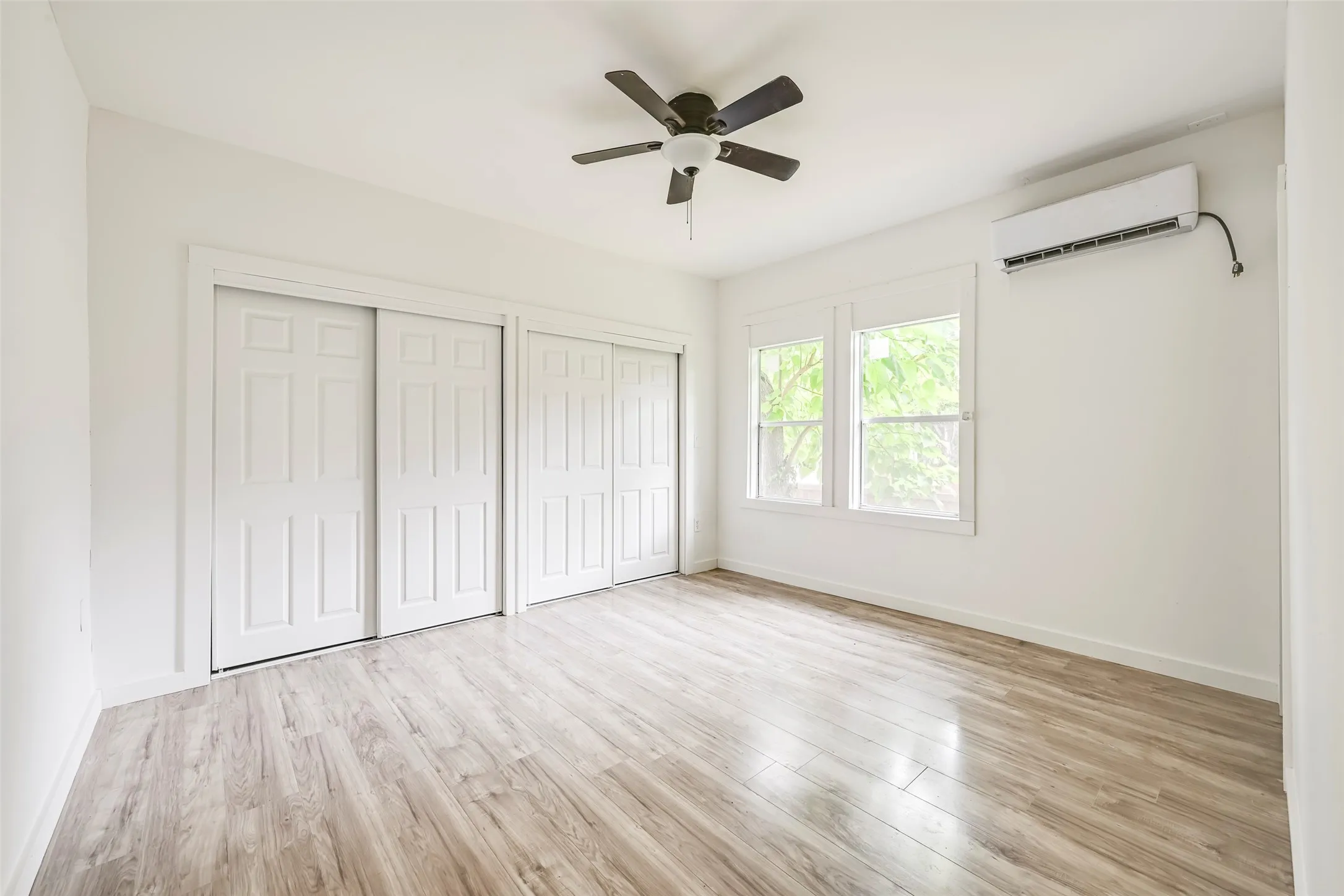 Bright room featuring wood-finish flooring, a ceiling fan, dual sliding closet doors, two windows, and a wall-mounted AC unit