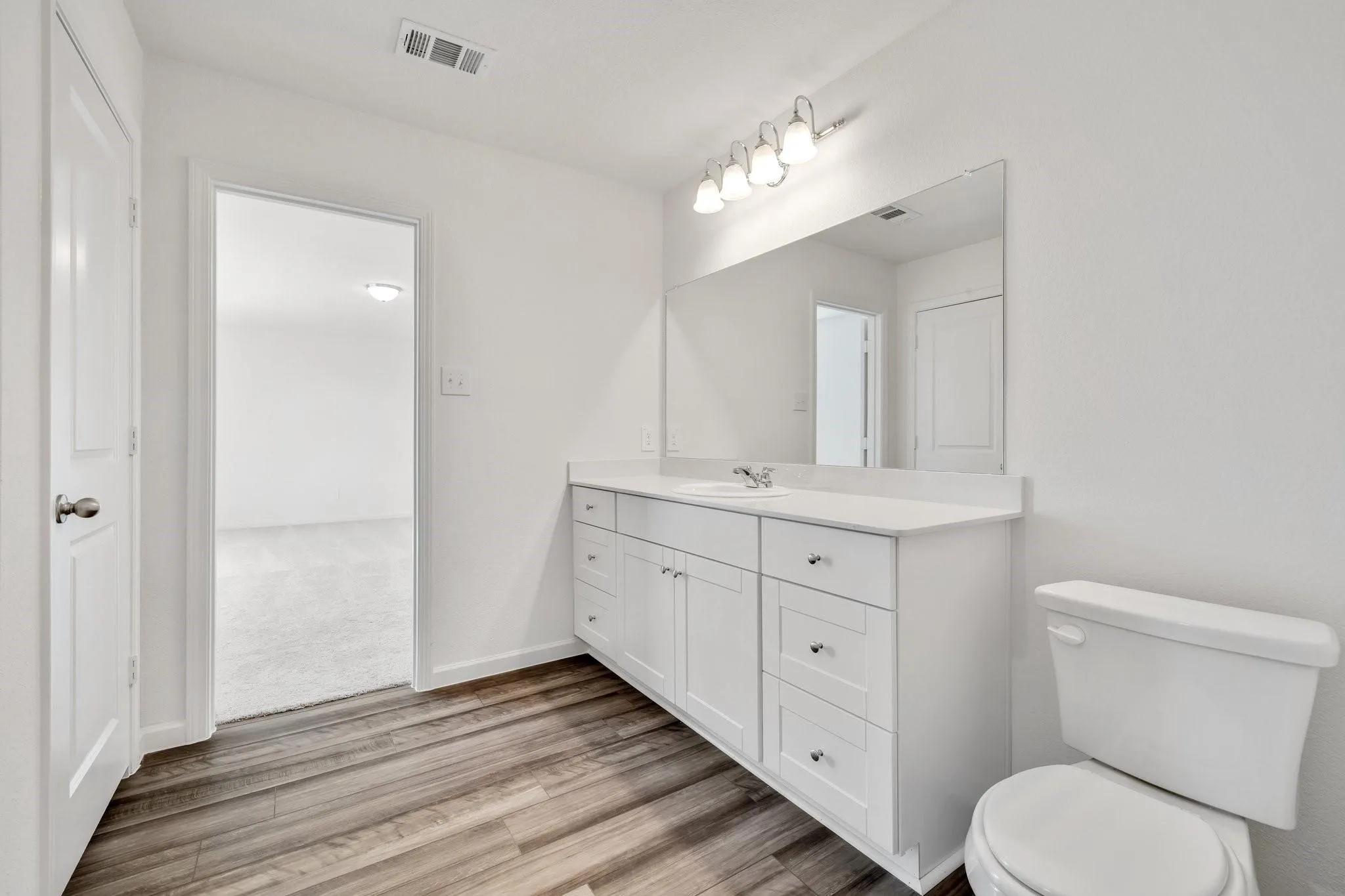 Spacious bathroom featuring wood-finish flooring, a white vanity with ample storage, a white countertop, and a large mirror with integrated lighting