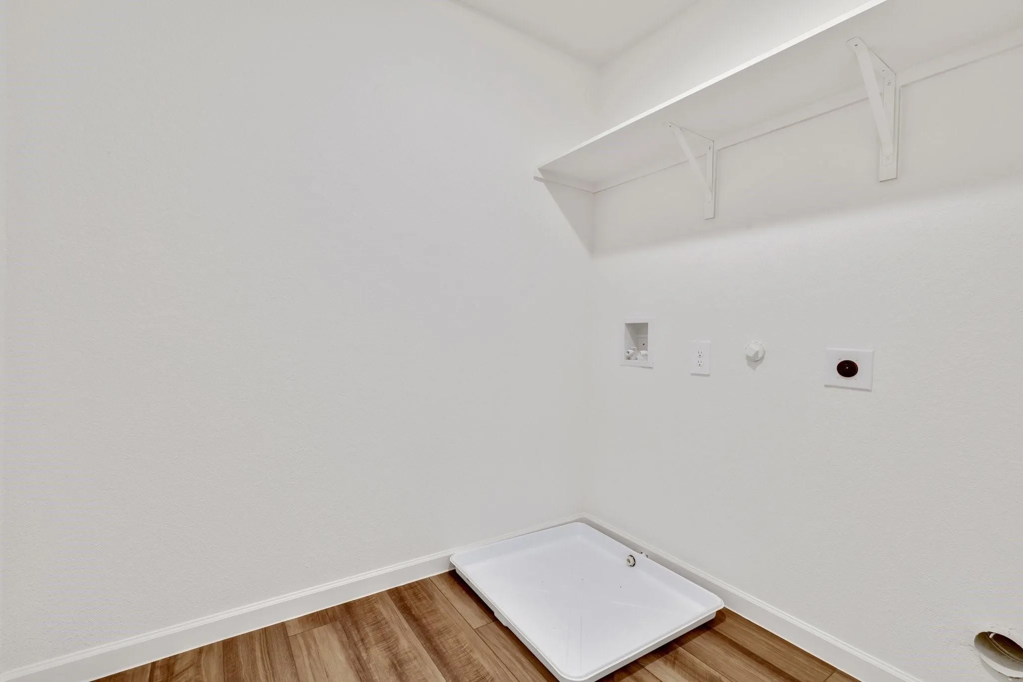 Dedicated laundry area featuring wood-finish flooring, a white wall shelf, a washing machine pan, and essential utility connections