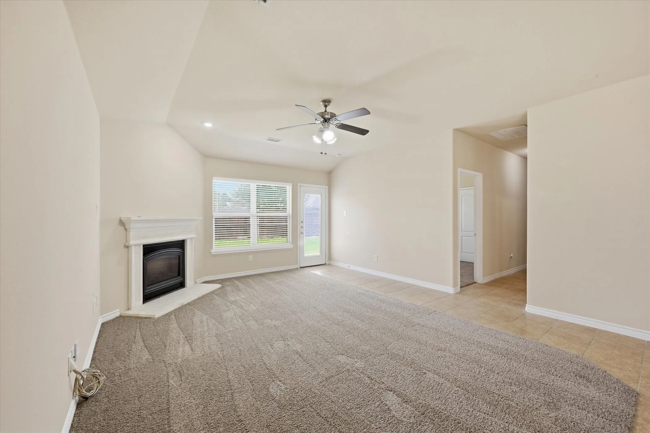 Spacious living area featuring neutral-toned carpeting, a corner fireplace with a white mantel, and a ceiling fan with integrated lighting