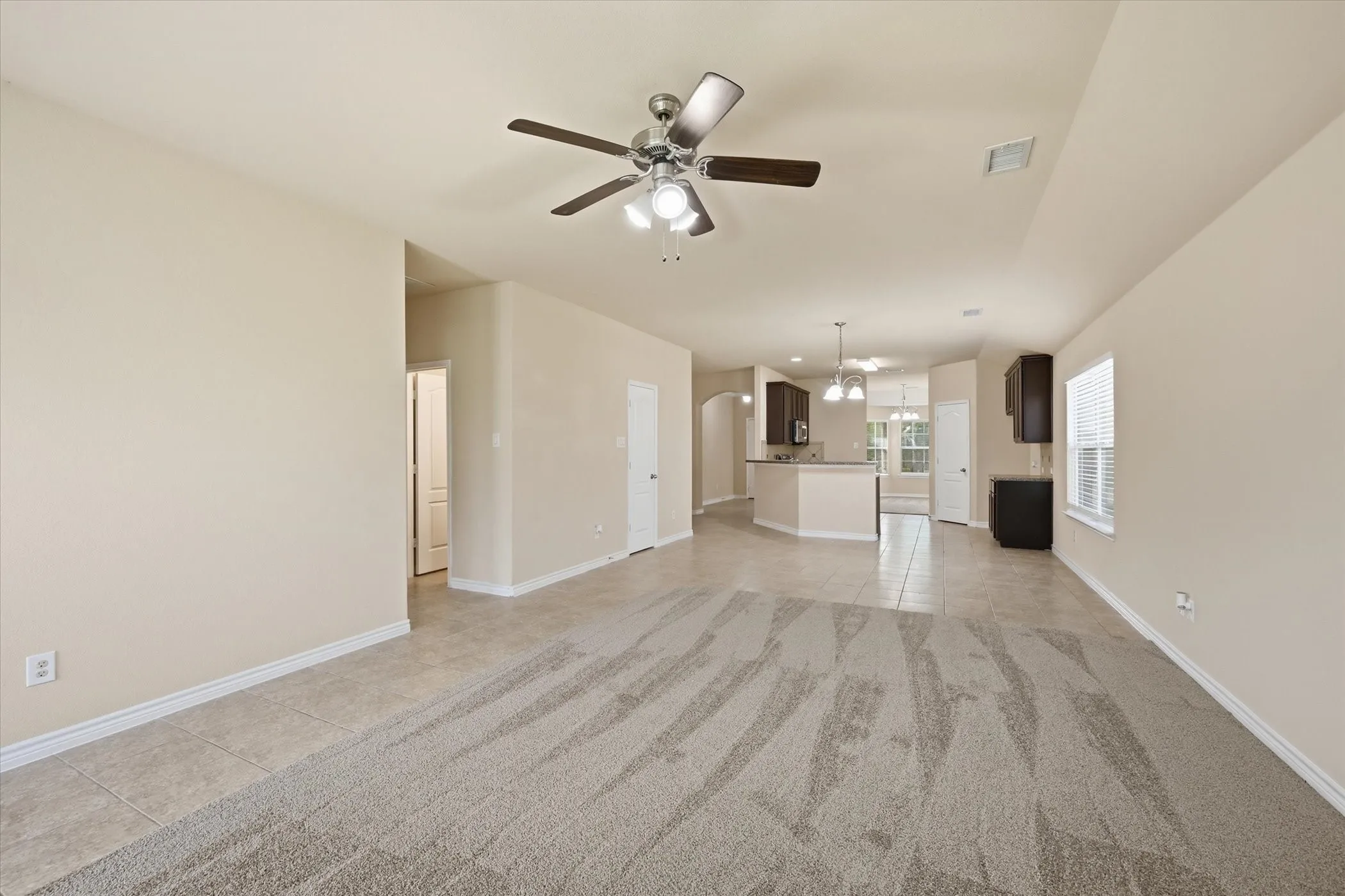 Spacious open-concept room featuring carpet and tile flooring, a ceiling fan with integrated lighting, and neutral wall tones