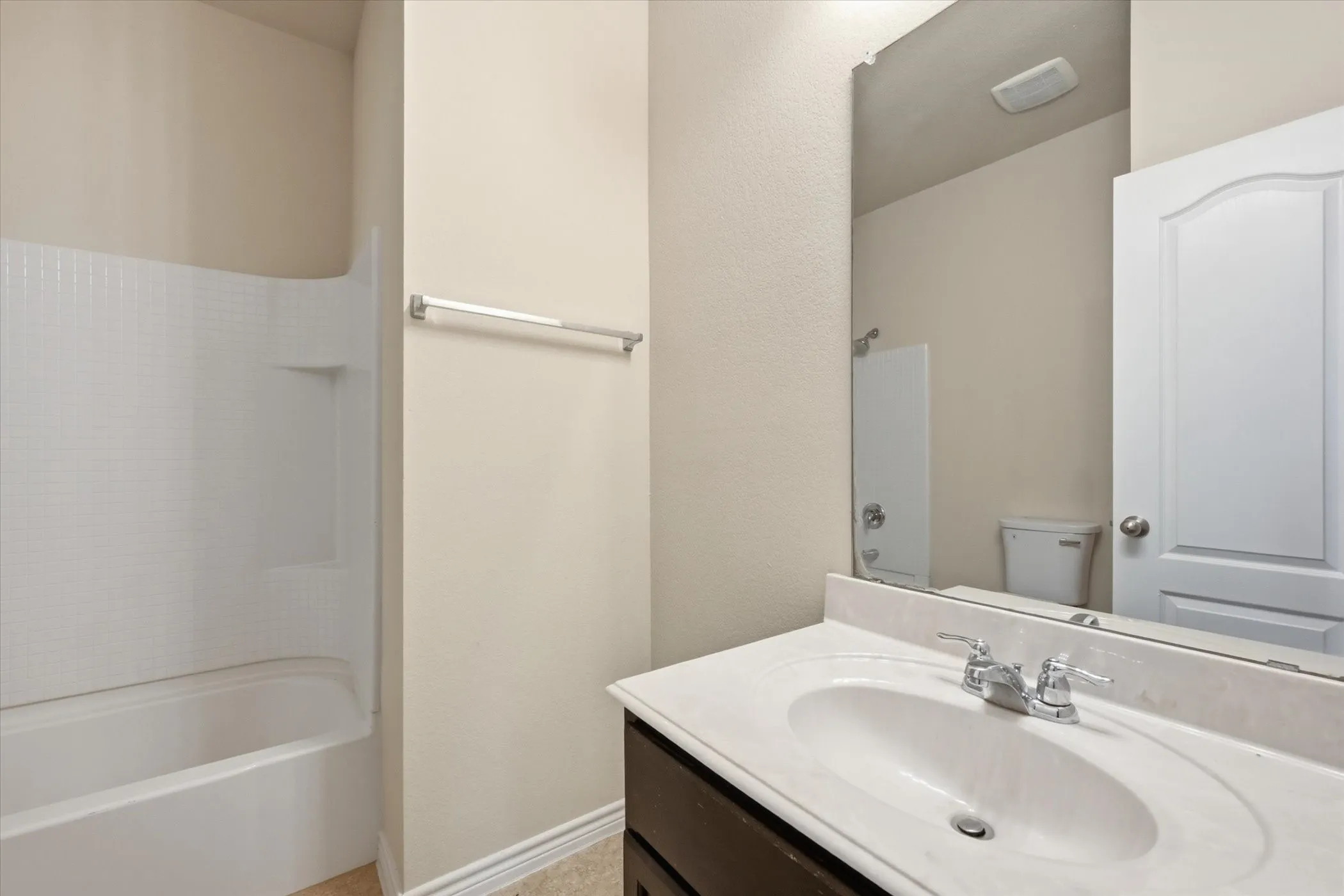 Bathroom featuring an integrated bathtub and shower with a tiled surround, a dark wood-finish vanity with an integrated sink, a light-toned countertop, a wall-mounted mirror, and a chrome faucet