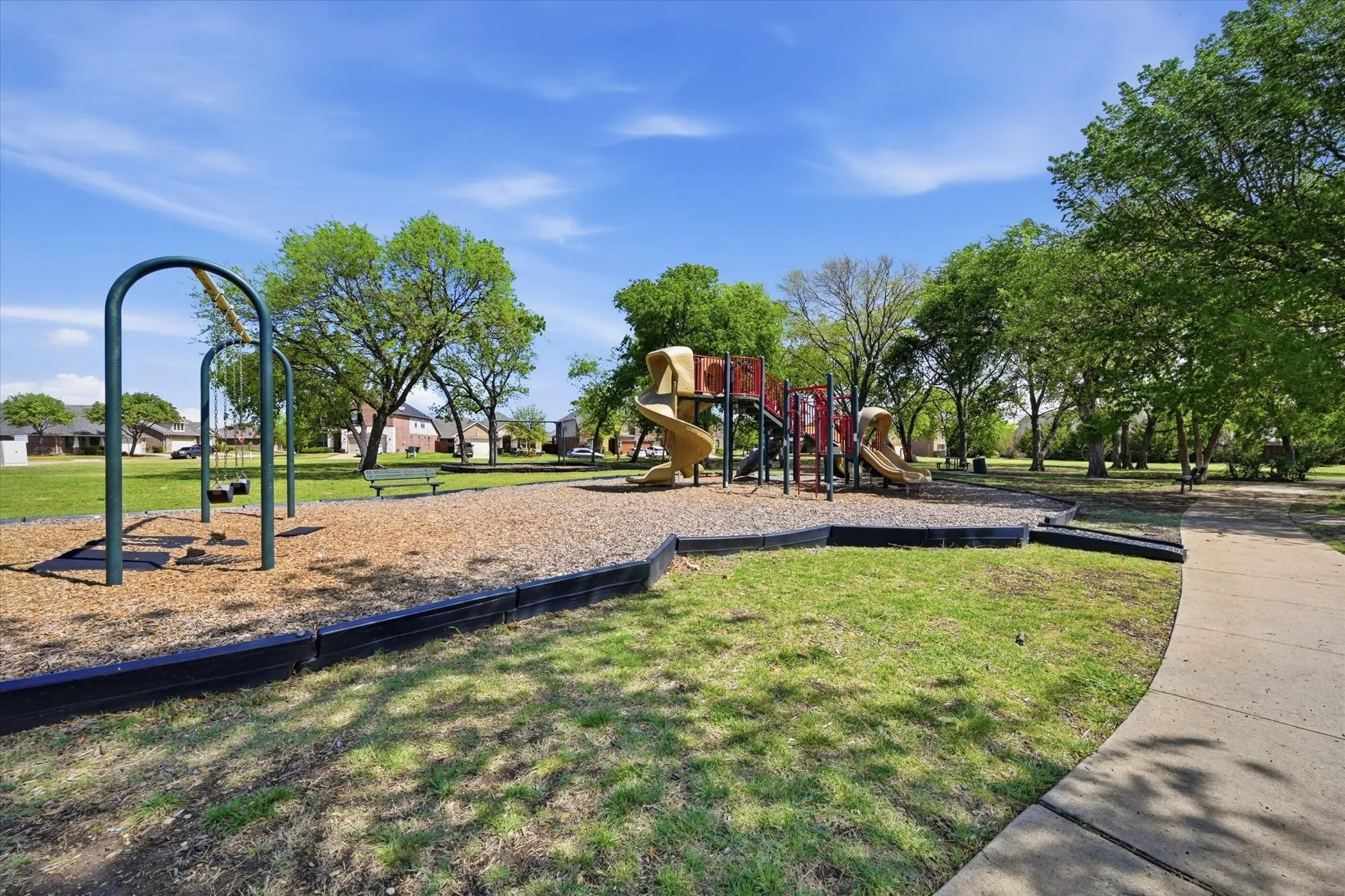 Community park featuring a playground with swings and slides on a wood chip surface, surrounded by grassy areas and mature trees