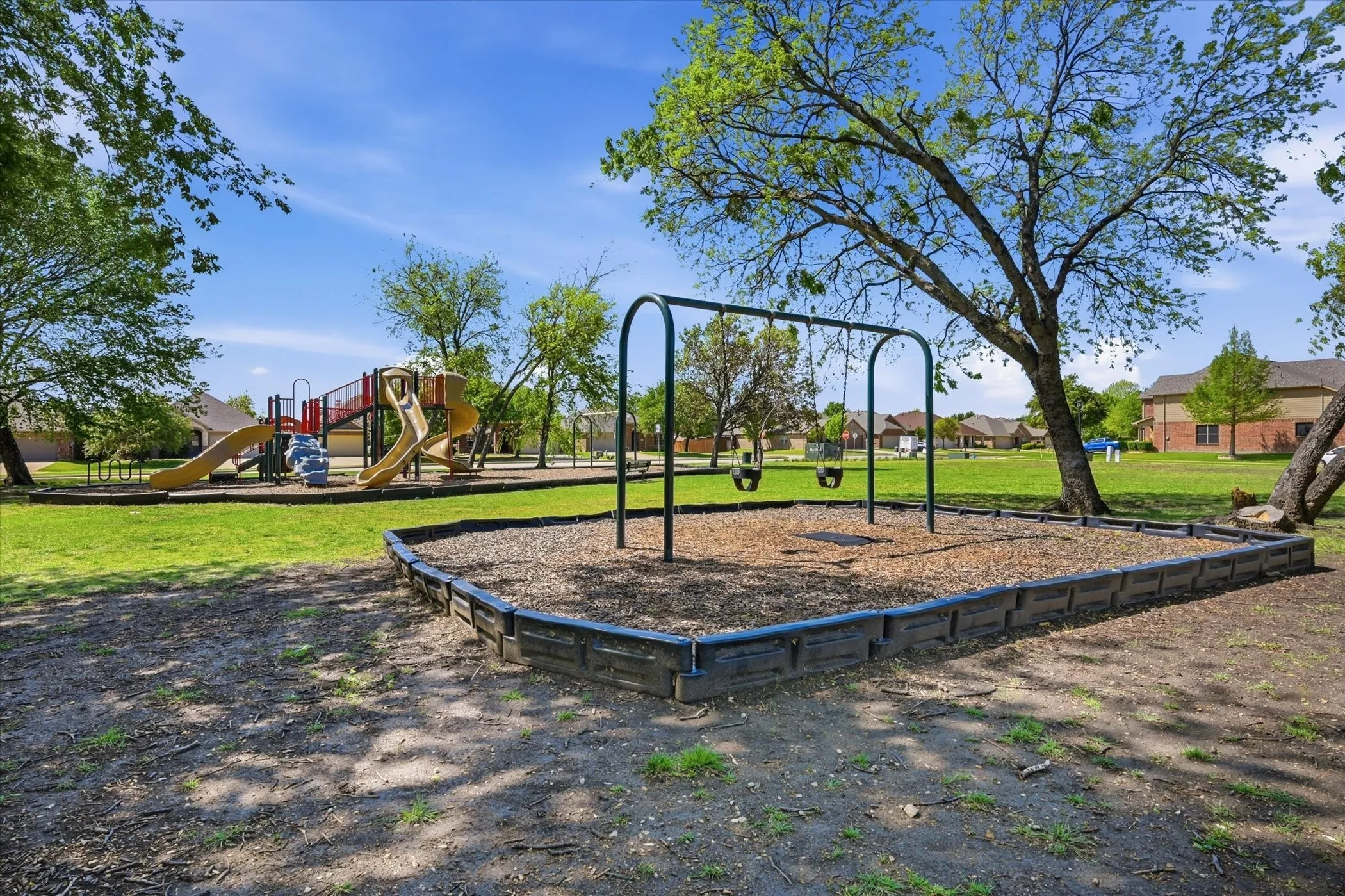 Community playground featuring a swingset with wood chip surfacing, a multi-level play structure with slides, and mature trees providing natural shade
