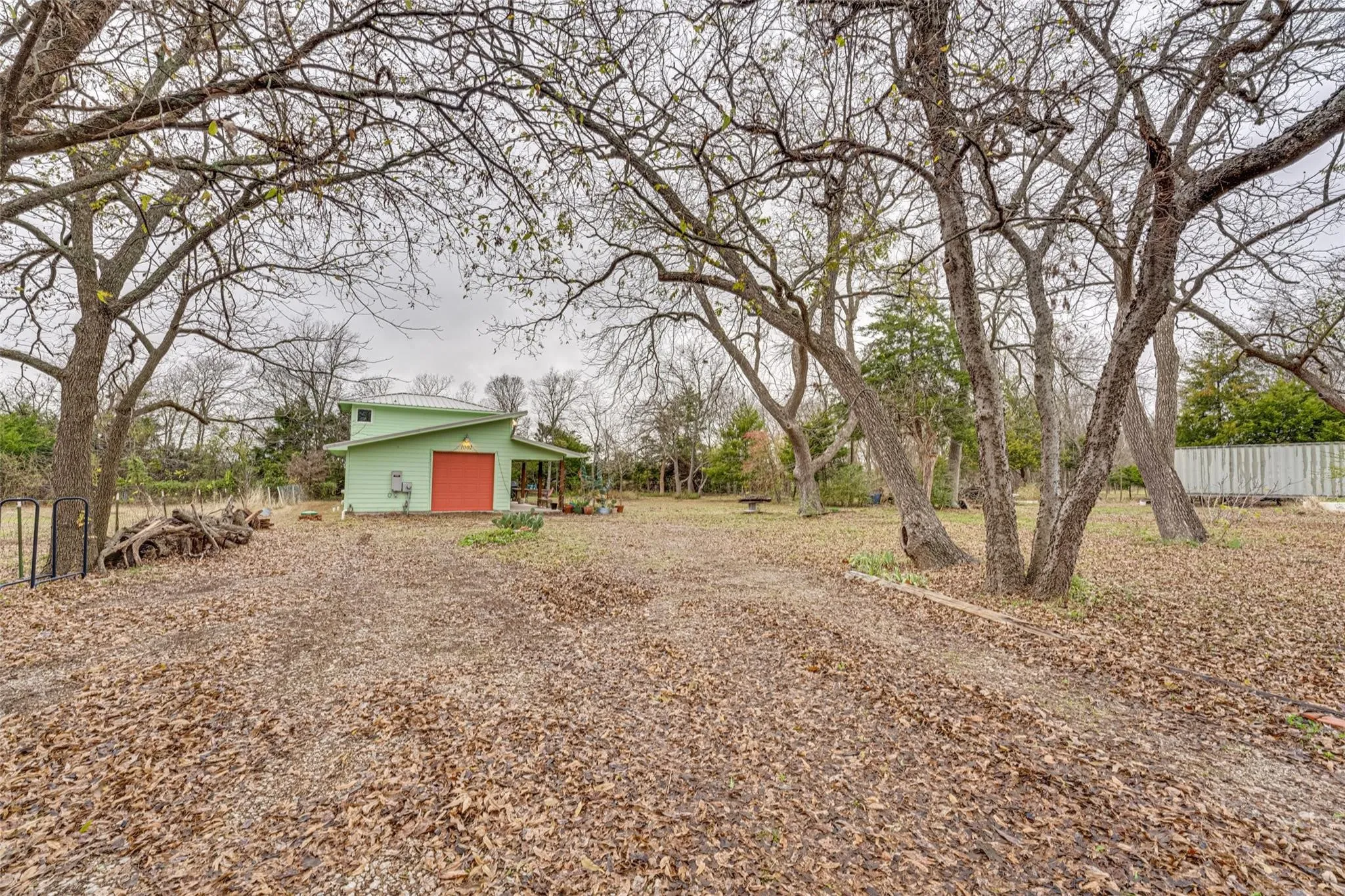 Expansive outdoor area featuring numerous mature trees and a detached structure with a pitched roof, light green siding, and a red roll-up door