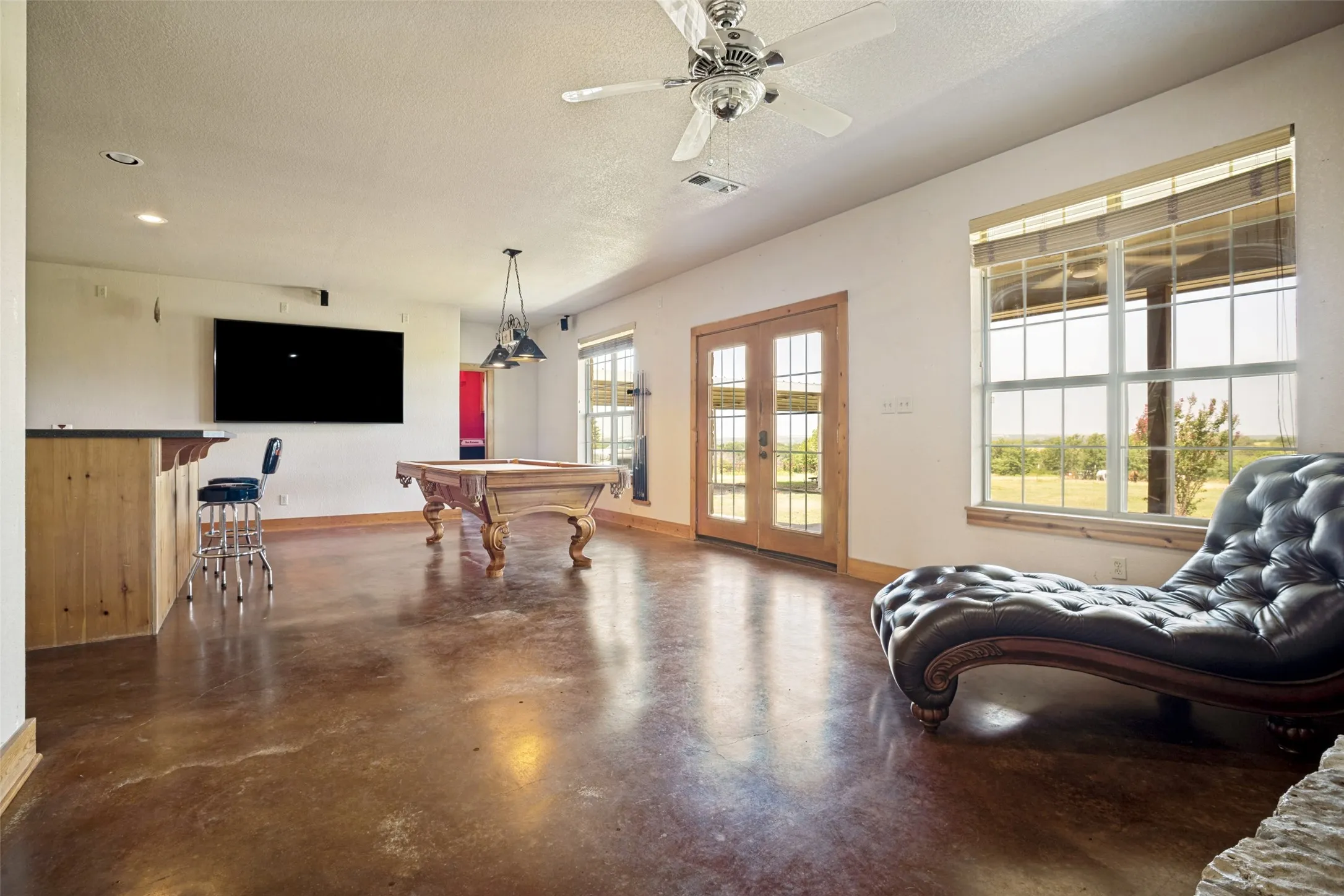 Spacious entertainment room featuring stained concrete flooring, a built-in bar, French doors, and large windows with blinds