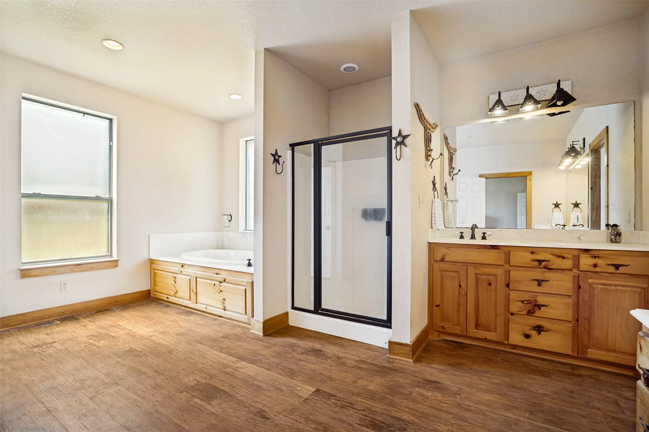 Spacious bathroom featuring wood-finish flooring, a soaking tub with a tiled surround and wood cabinetry, a glass-enclosed shower, and a double vanity with a large mirror and three-light fixture