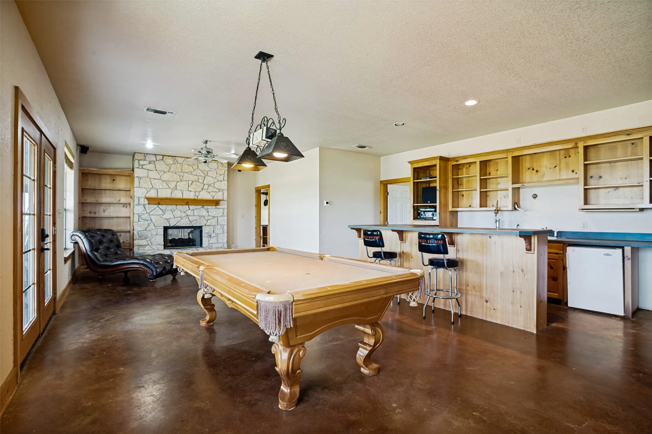 Spacious entertainment room featuring a stone-faced fireplace with wood mantel, built-in shelving, stained concrete flooring, and a built-in bar with a countertop