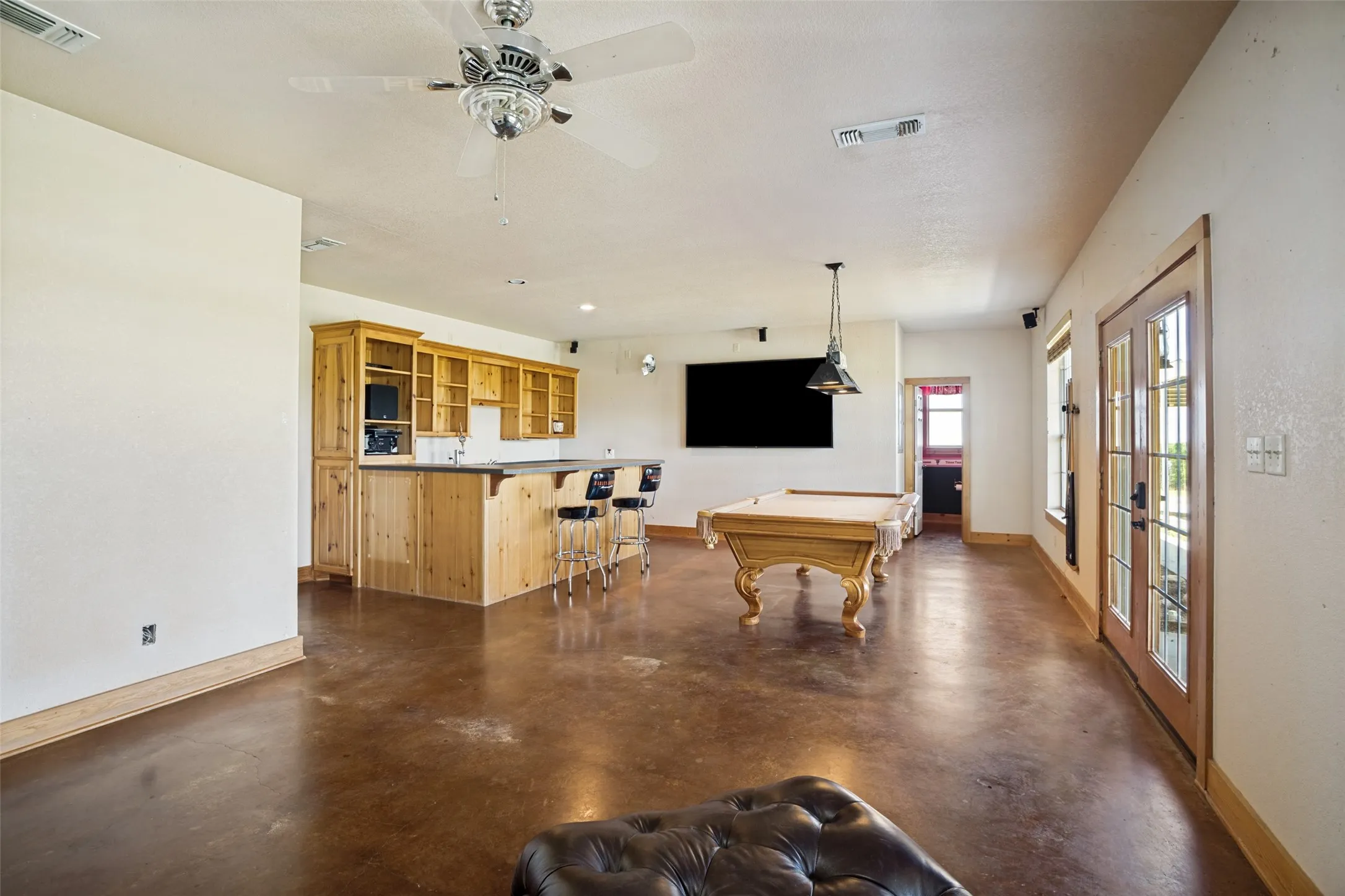 Spacious entertainment area featuring stained concrete flooring, a custom wood wet bar with built-in cabinetry, dual French doors with glass panels, and a ceiling fan with light fixture