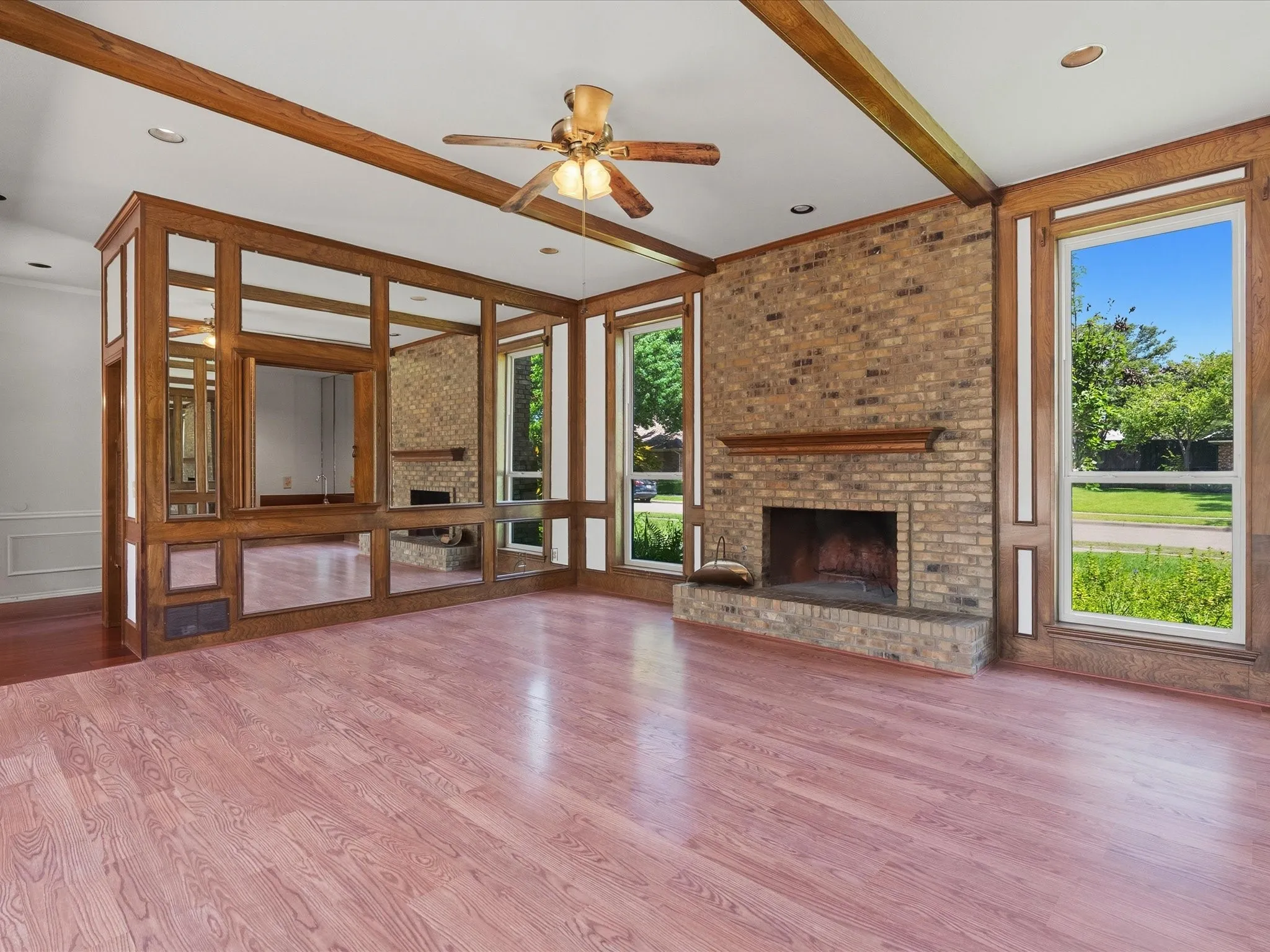 Living space featuring wood-finish flooring, a brick fireplace with a wood mantle, an exposed beam ceiling, a mirrored accent wall with wood trim, and large windows