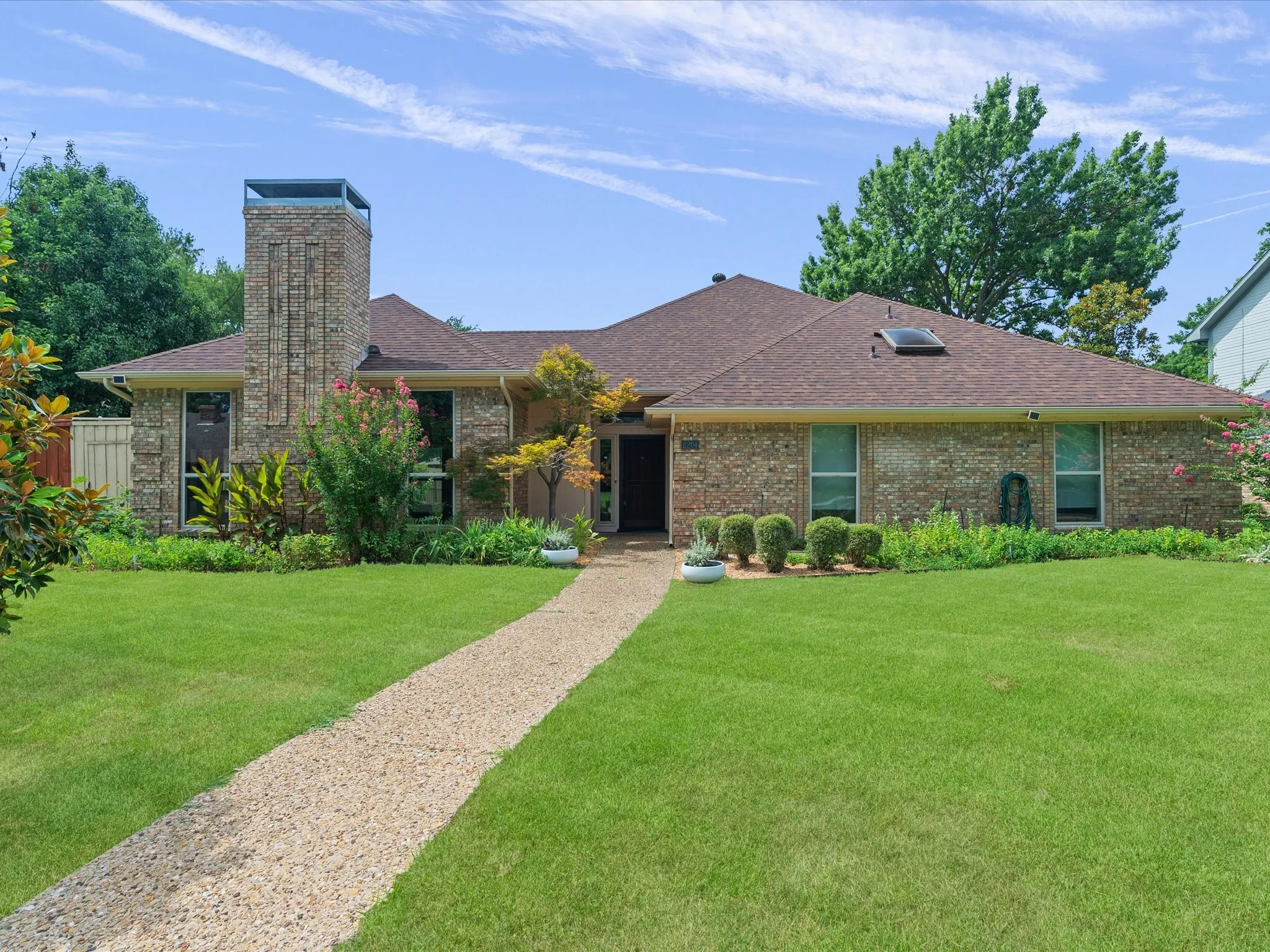 Brick facade residence featuring a prominent stone chimney, a warm brown shingle roof, a skylight, a gravel pathway, and mature landscaping
