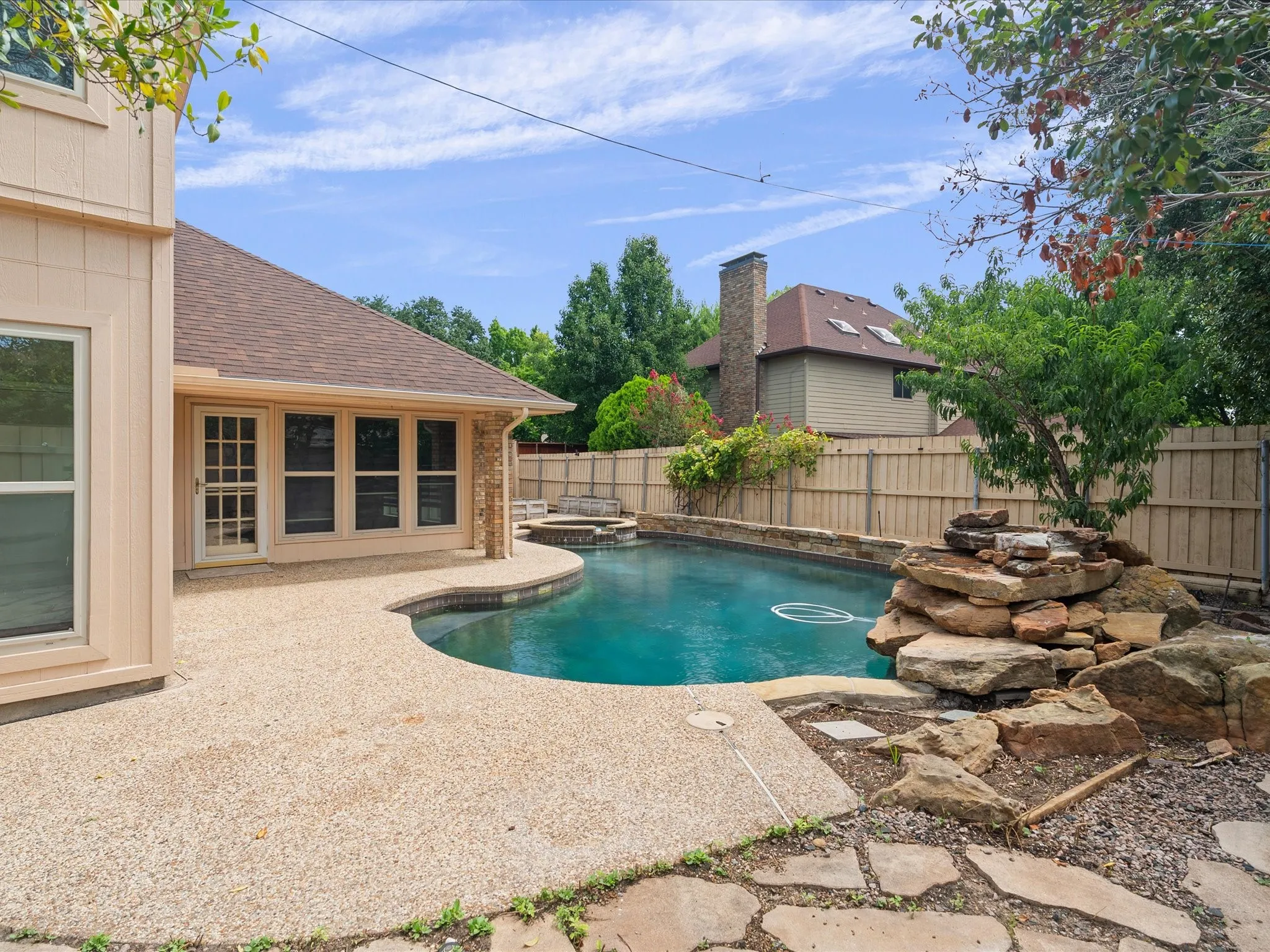 Curved swimming pool featuring an integrated spa, rock waterfall, and a surrounding aggregate patio