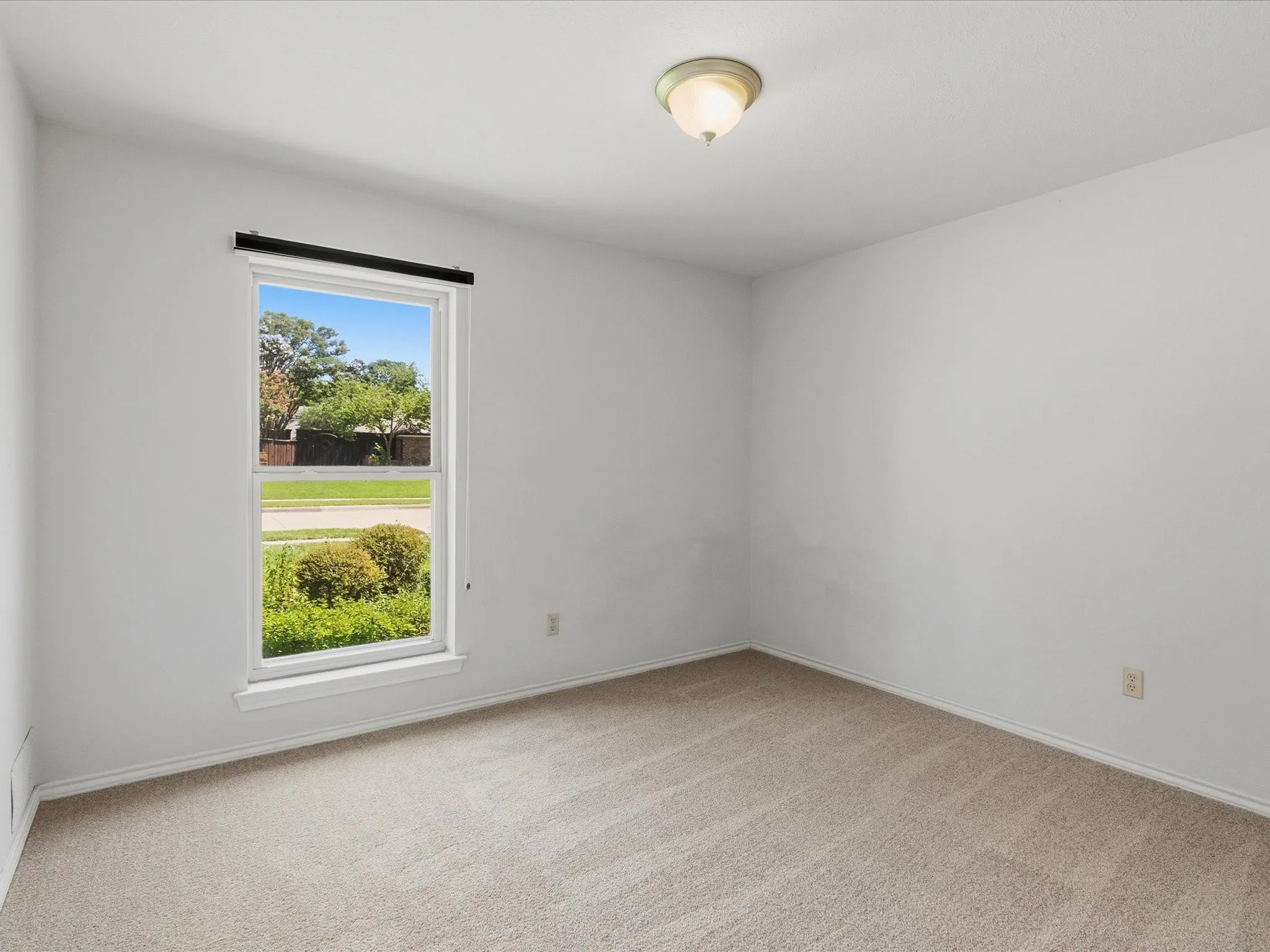 Bright interior space featuring a window with a roller shade, light-colored carpeting, white baseboards, and a semi-flush mount ceiling light fixture