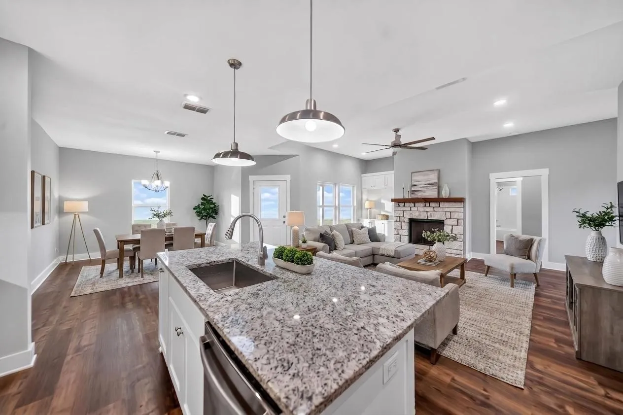 Open-concept living space featuring a central kitchen island with a stainless steel sink, wood-finish flooring, a stone fireplace with a wood mantel, vaulted ceilings, and recessed lighting. Virtually staged.