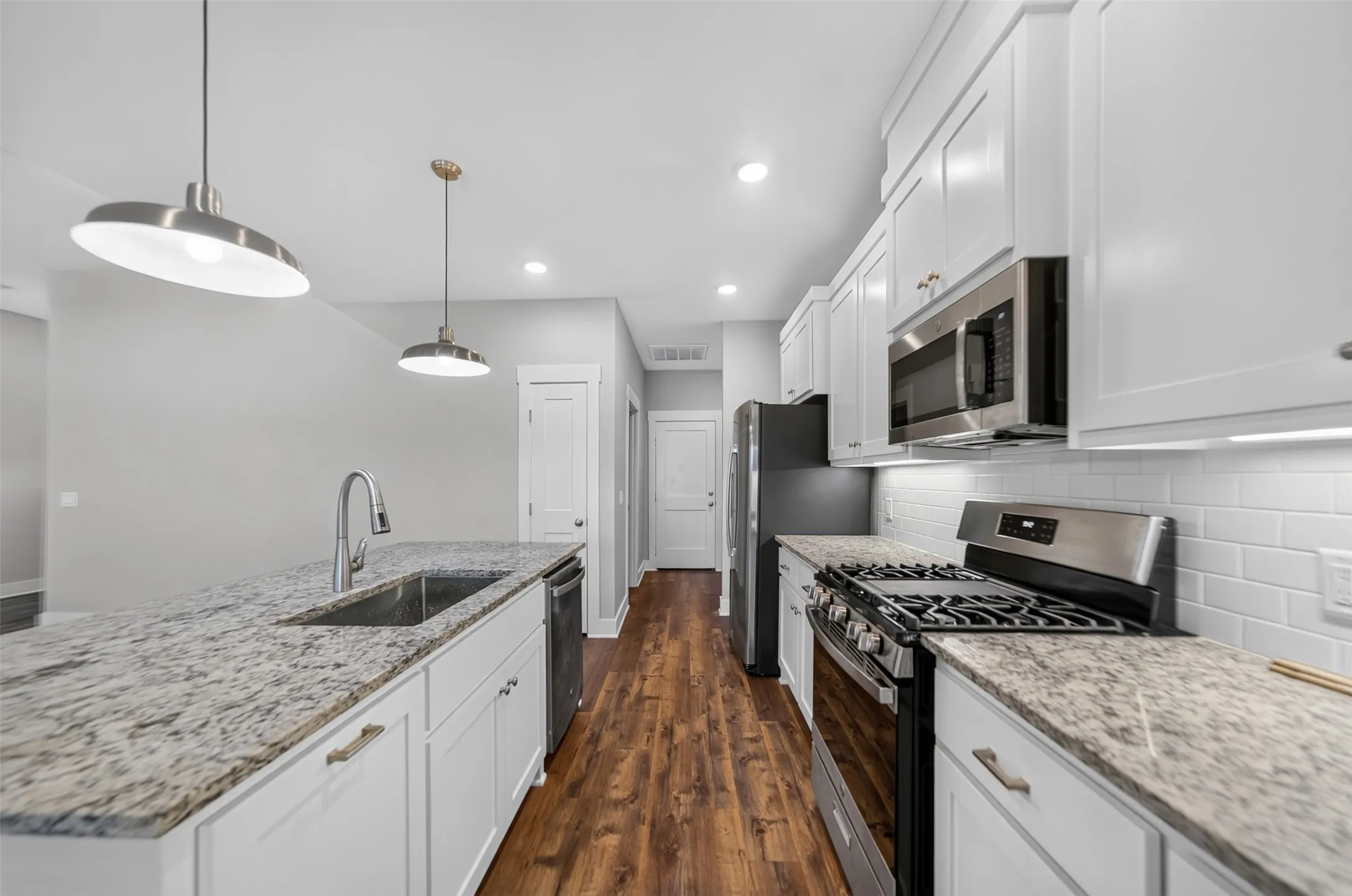 Kitchen featuring a center island with sink and pendant lighting, white shaker cabinetry, stainless steel appliances, speckled countertops, and wood-finish flooring