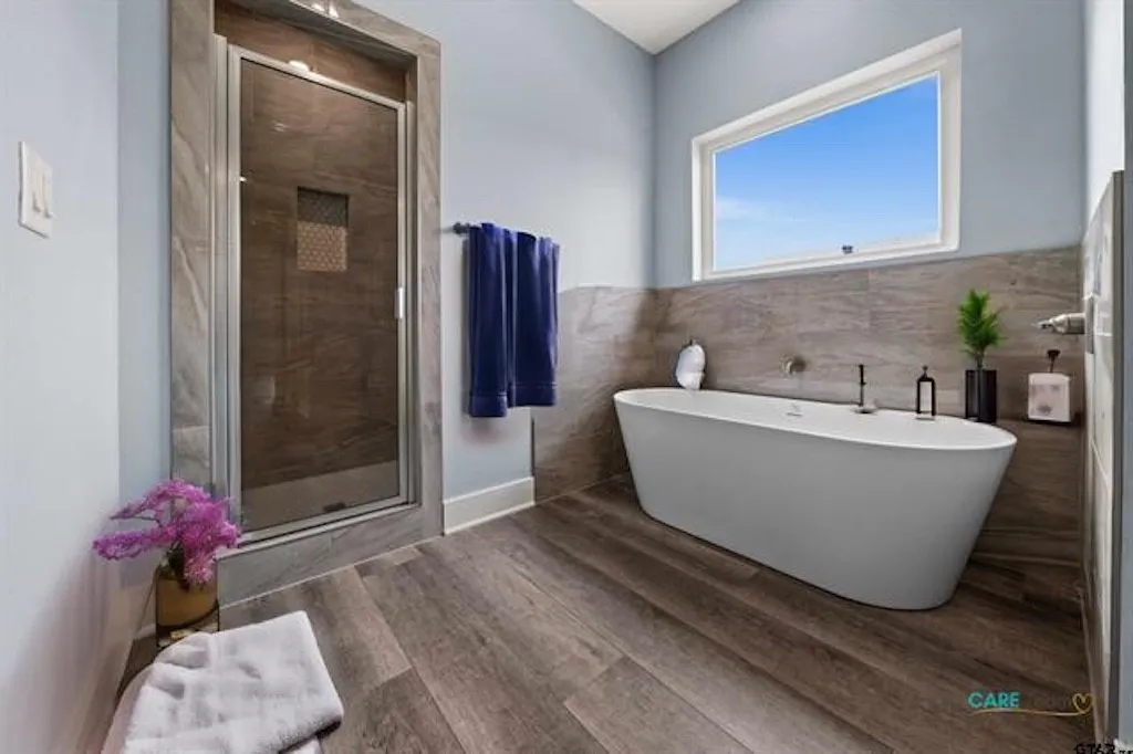 Freestanding soaking tub with floor-mounted faucet, complemented by wood-finish flooring and full-height wall tiling. Virtually staged.