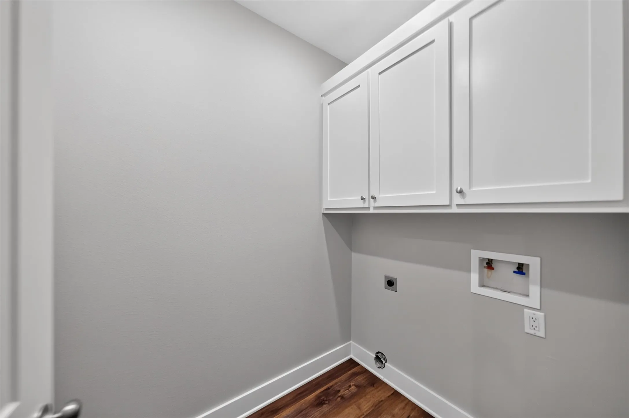 Laundry area featuring wood-finish flooring, white baseboards, and overhead white shaker-style cabinetry with brushed nickel hardware