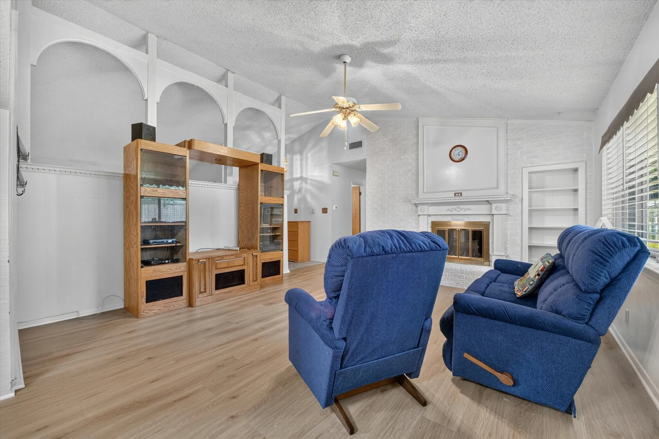 Living area featuring a fireplace with a decorative mantel, built-in shelving, wood-finish flooring, a ceiling fan, and window blinds