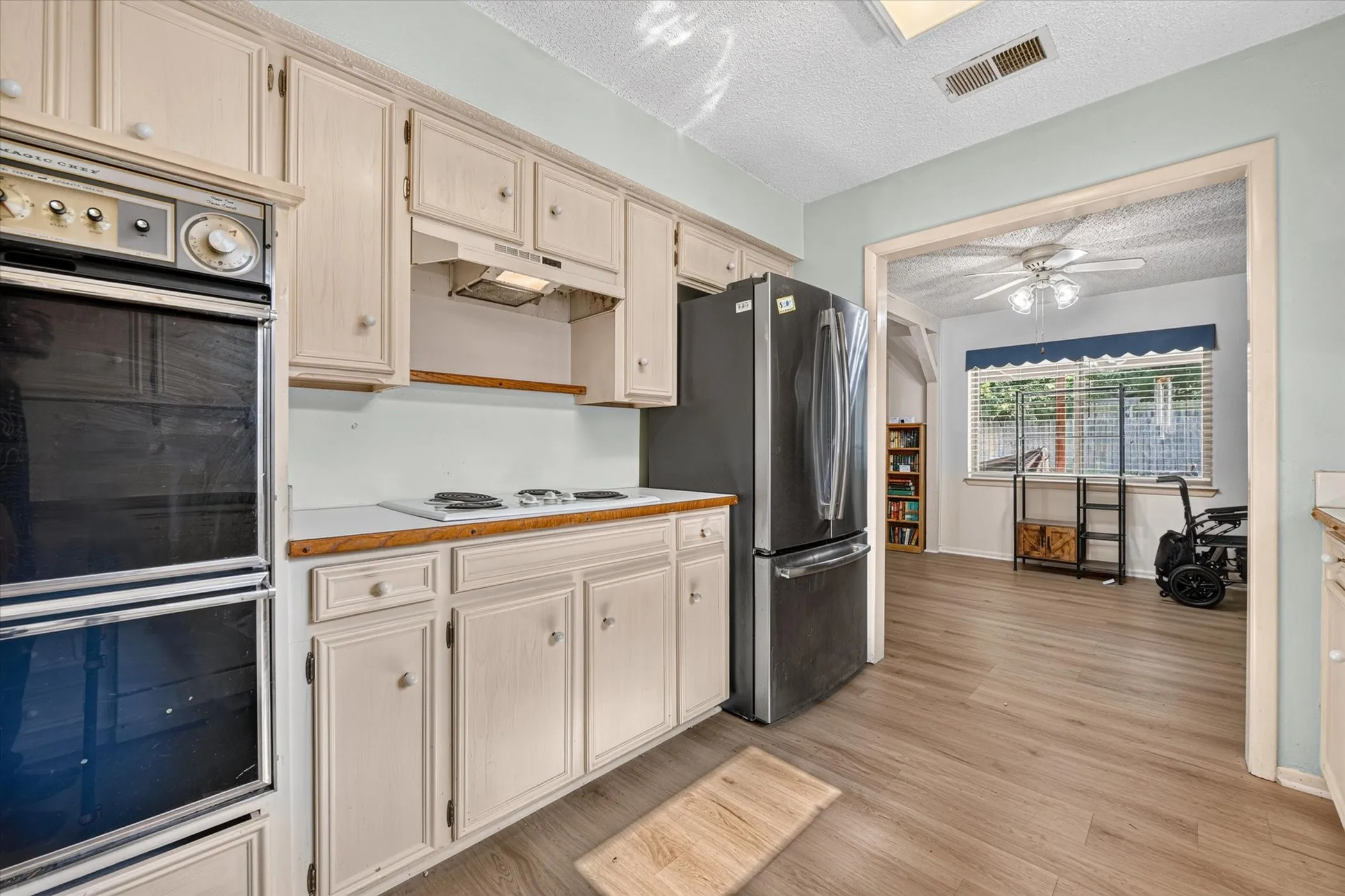 Kitchen featuring wood-finish flooring, light-toned cabinetry, double wall ovens, a black refrigerator, and a built-in cooktop with a range hood