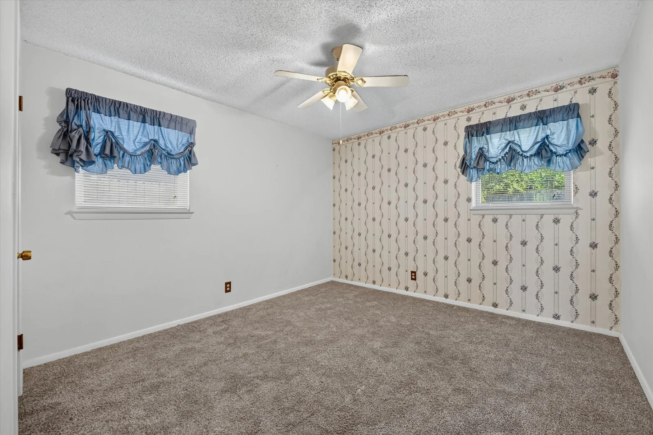 Carpeted room featuring a ceiling fan with integrated lighting, two windows with blinds, and one wall adorned with patterned wallpaper