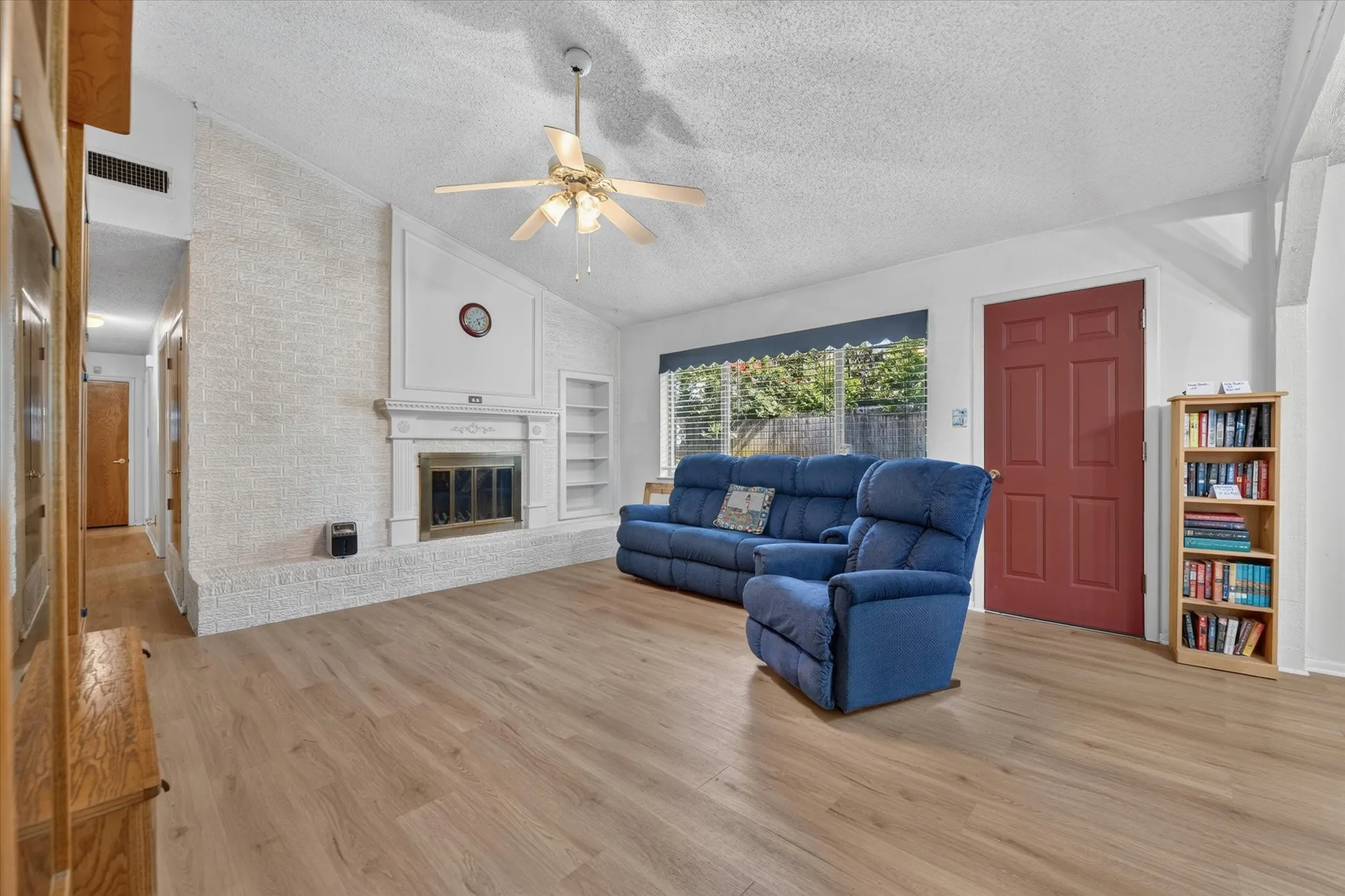 Spacious living area featuring light wood-finish flooring, a white brick fireplace with mantel, built-in shelving, a vaulted ceiling, and a ceiling fan