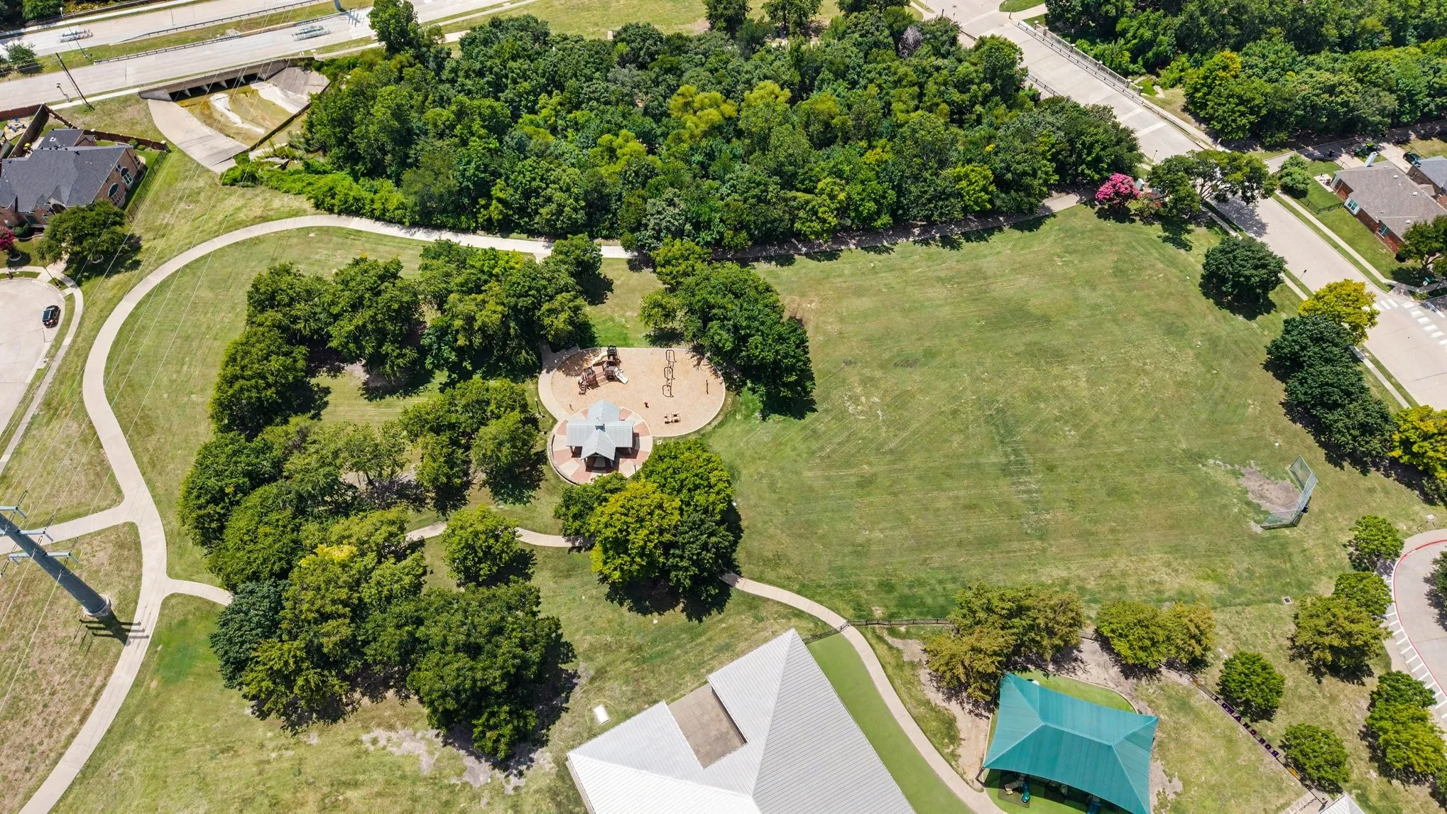 Expansive park grounds featuring open green spaces, mature trees, a playground with a covered seating area, paved walking paths, and a dedicated sporting area