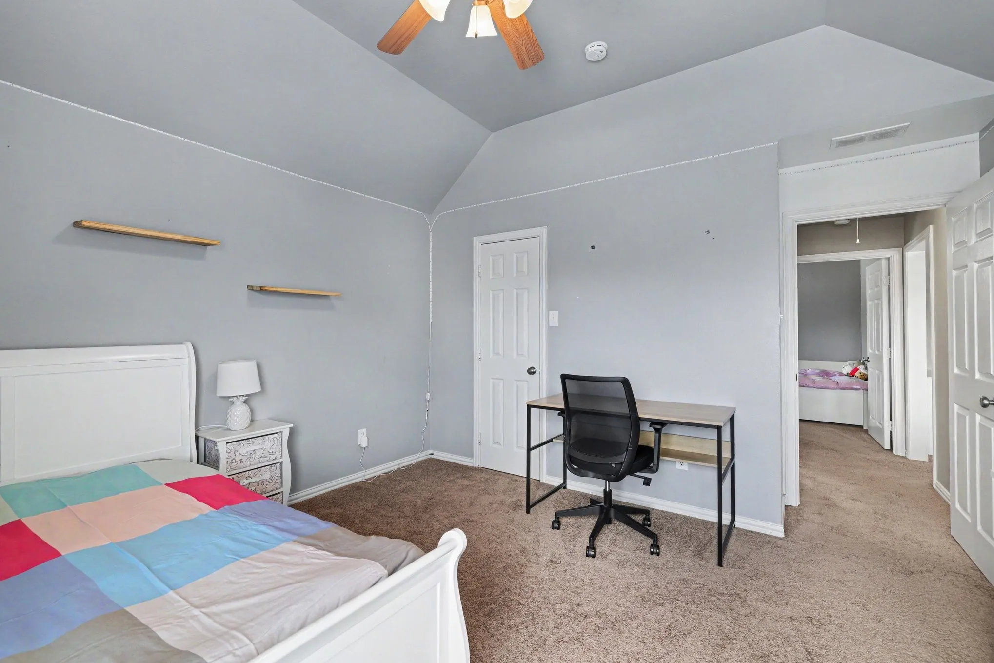Carpeted bedroom with a vaulted ceiling, ceiling fan, two wall-mounted shelves, and a white paneled door