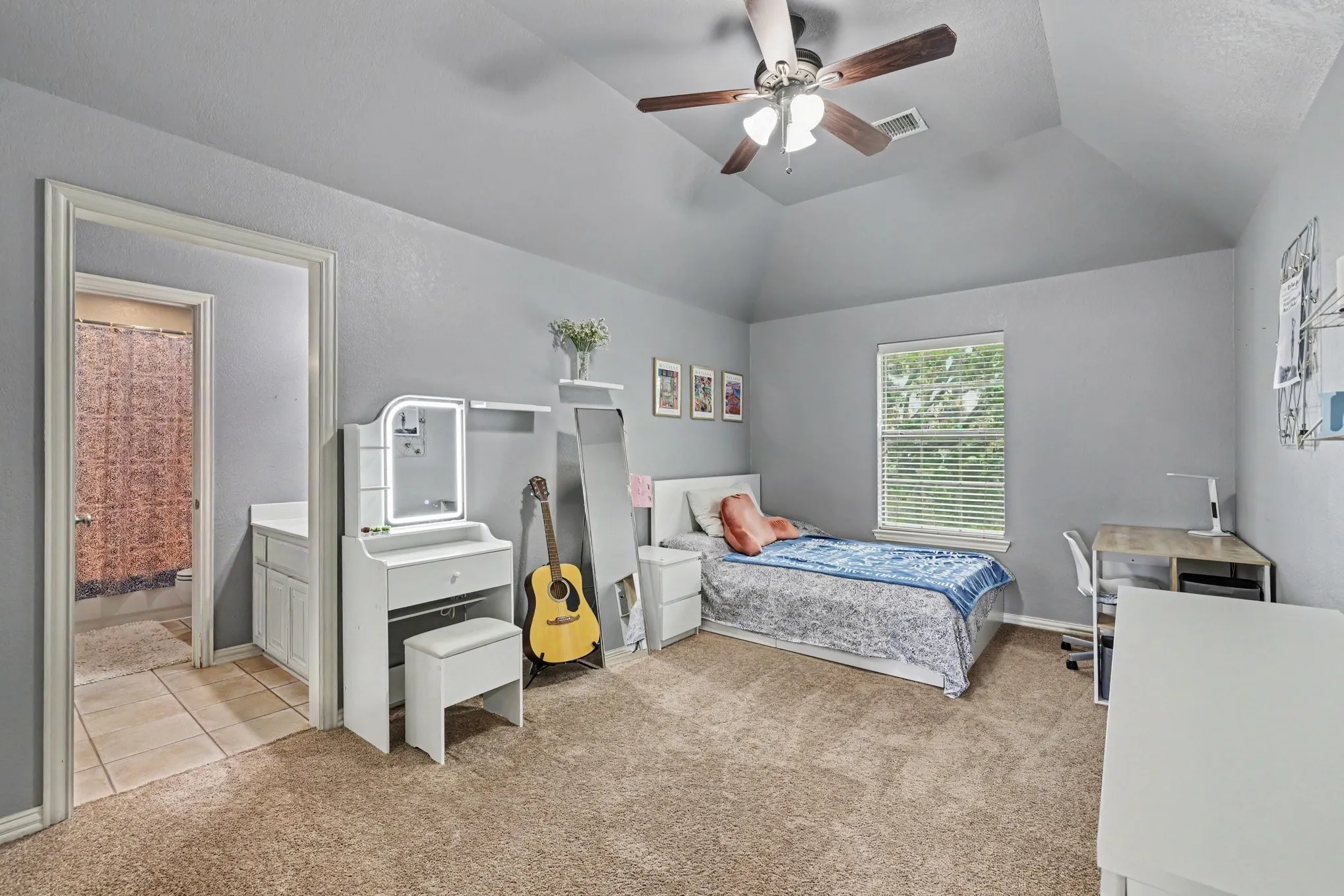 Carpeted room featuring a window with blinds, a ceiling fan, and an en-suite bathroom entrance with tile flooring and a built-in vanity