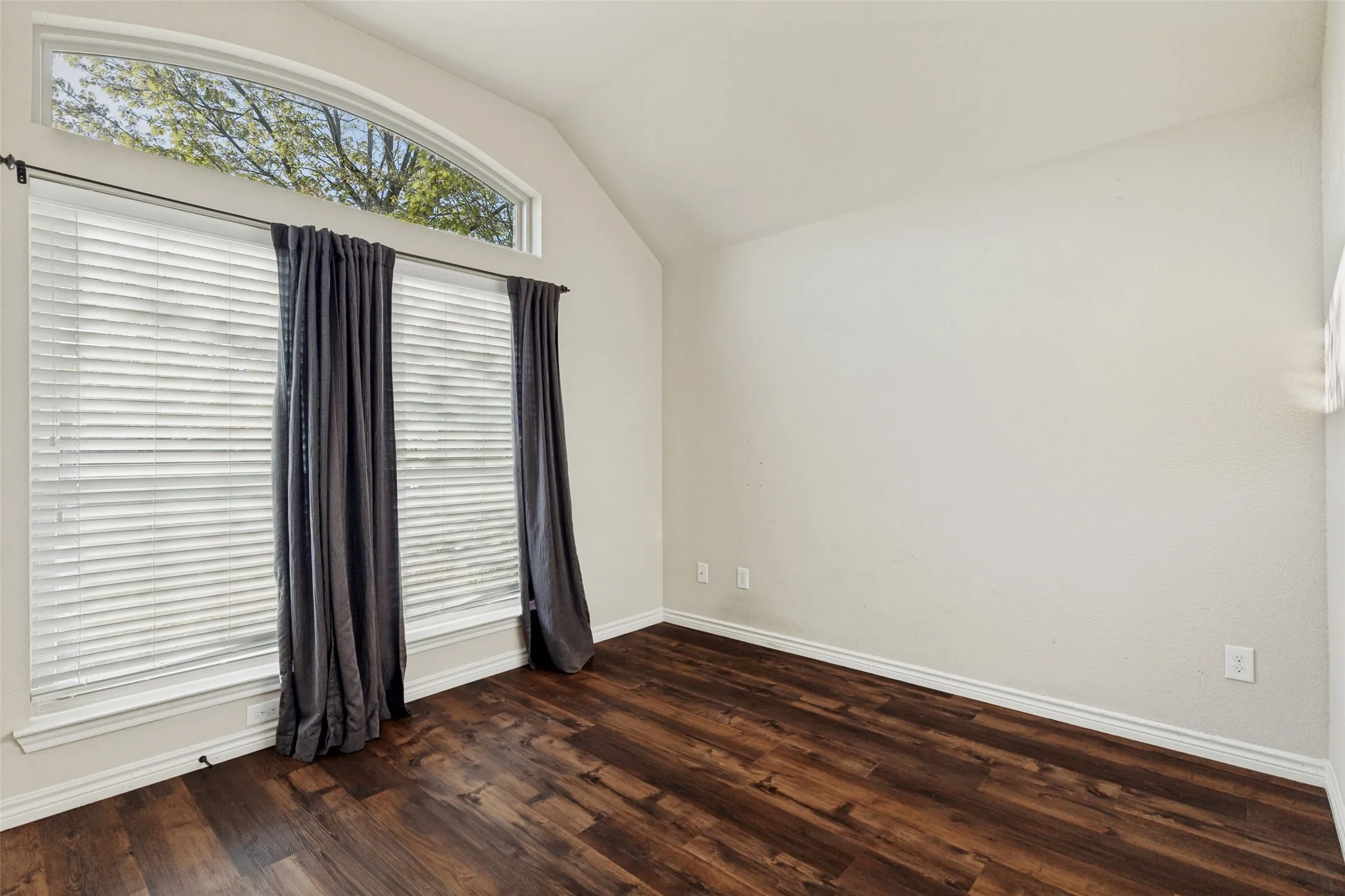 Vaulted ceiling room featuring wood-finish flooring, a large window with blinds, and an arched transom window