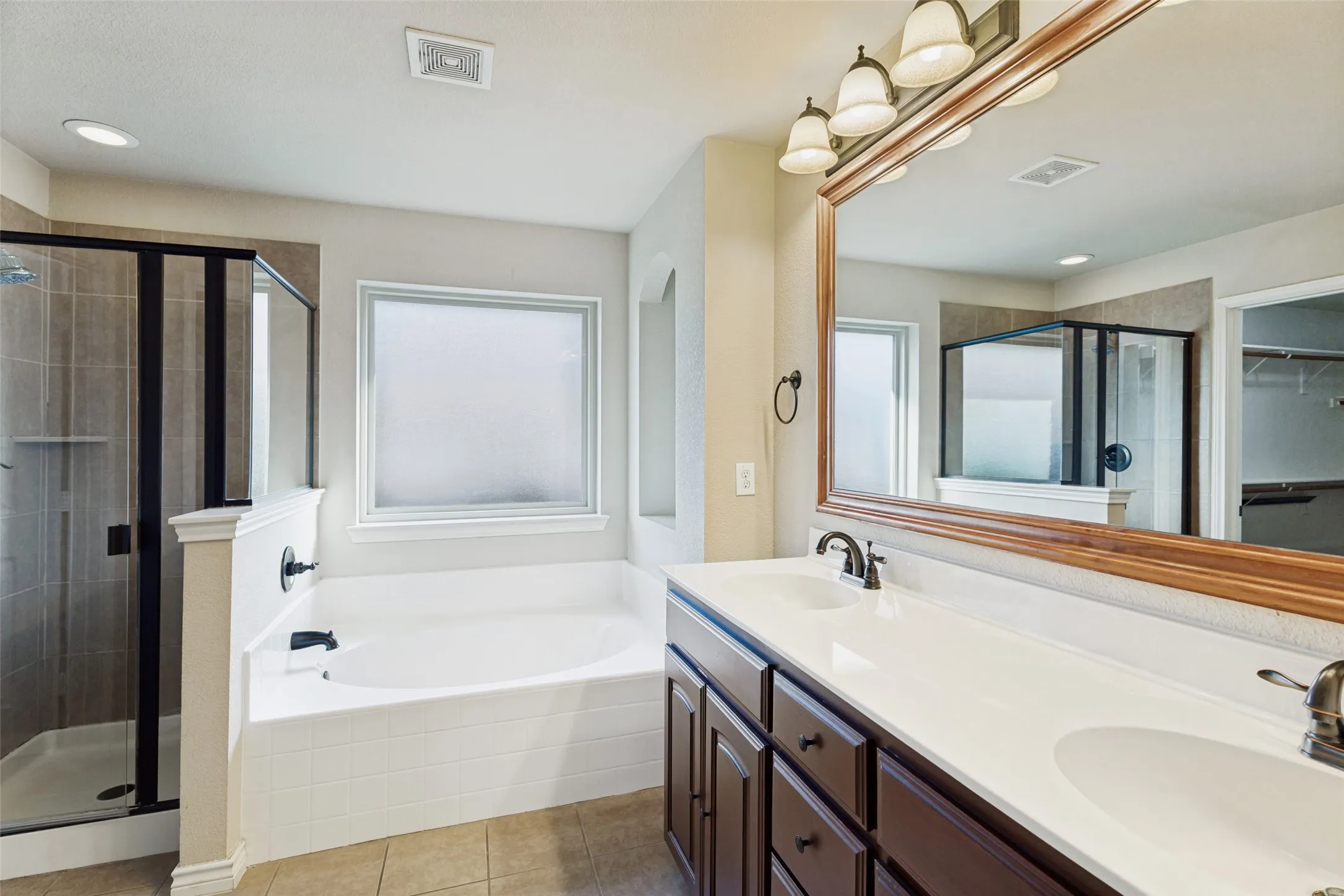 Bathroom featuring a dual vanity, large soaking tub, separate glass-enclosed shower, wood-finish cabinetry, and a framed mirror