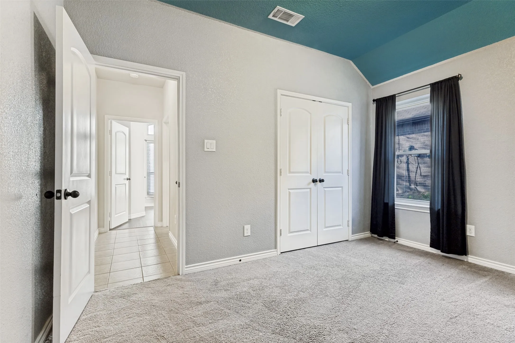 Vaulted ceiling with a teal accent, light gray walls, plush carpet flooring, a window with black curtains, and double-door closet