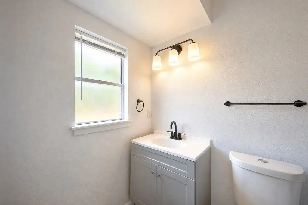 Bathroom featuring a single window, a gray vanity with an integrated sink, a dark finish faucet, a three-light fixture, and a white toilet
