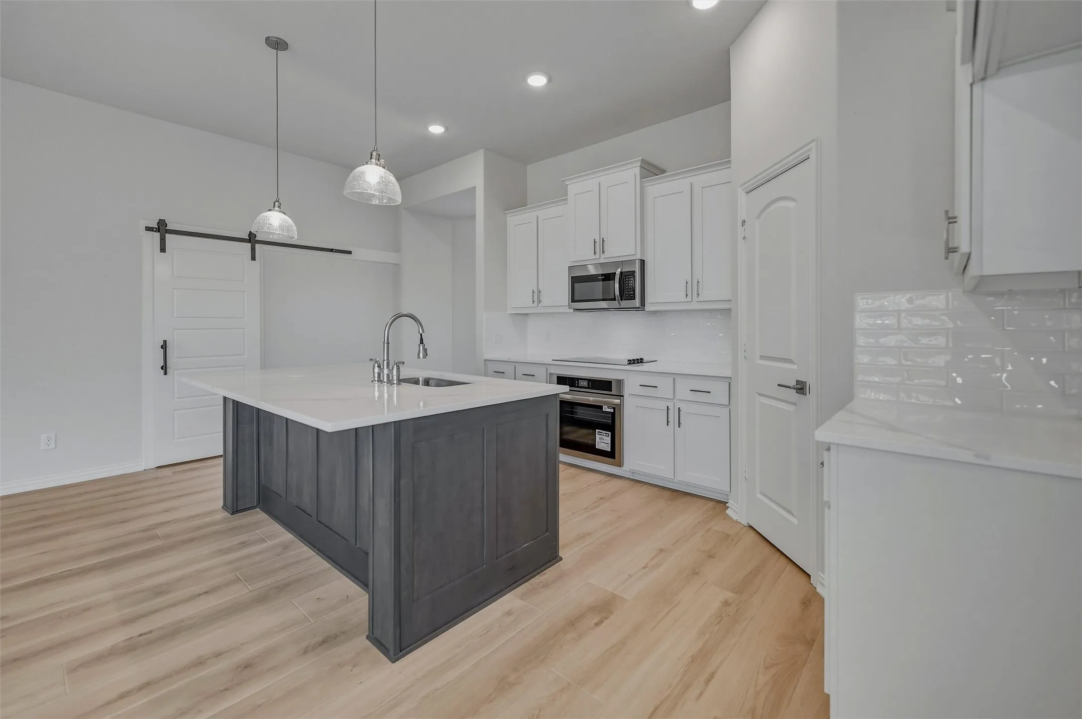 Kitchen featuring a central island with a sink, white cabinetry, integrated stainless steel appliances, light wood-finish flooring, and a white subway tile backsplash