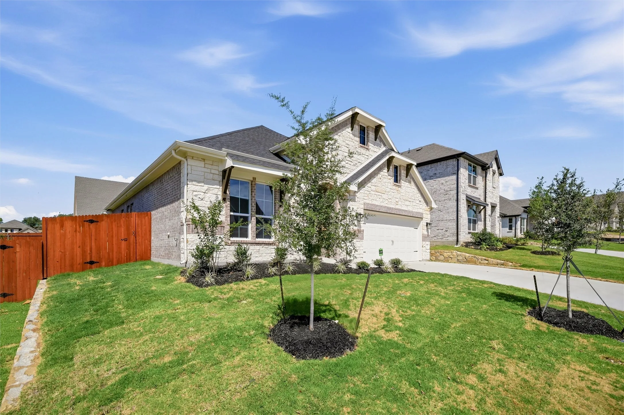 Stone and brick facade, dark shingle roof, attached garage, wood privacy fence, and paved driveway