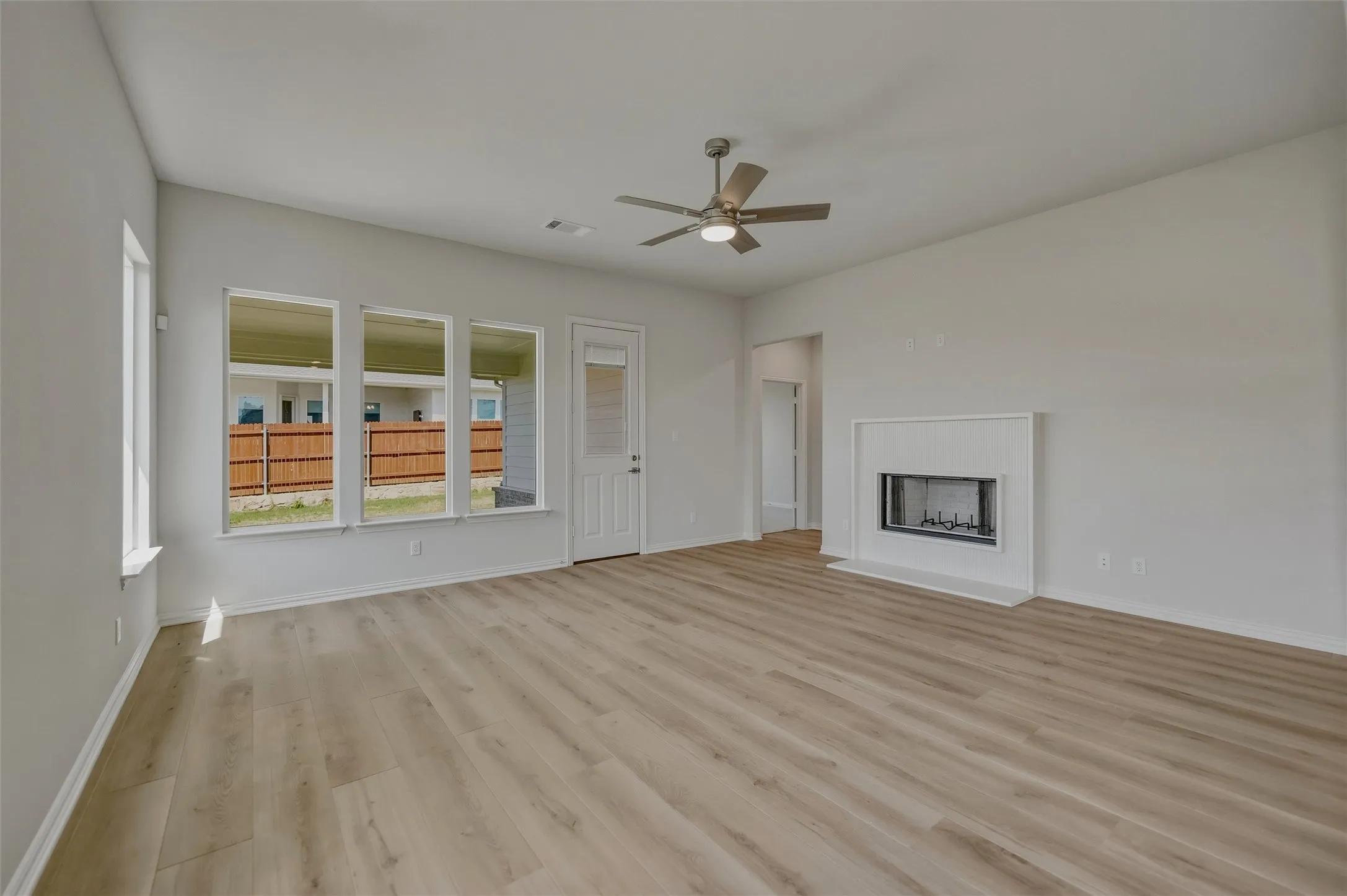 Expansive living space featuring wood-finish flooring, a contemporary ceiling fan, multiple windows for natural light, a white paneled door with glass insert, and a modern fireplace with a white mantel